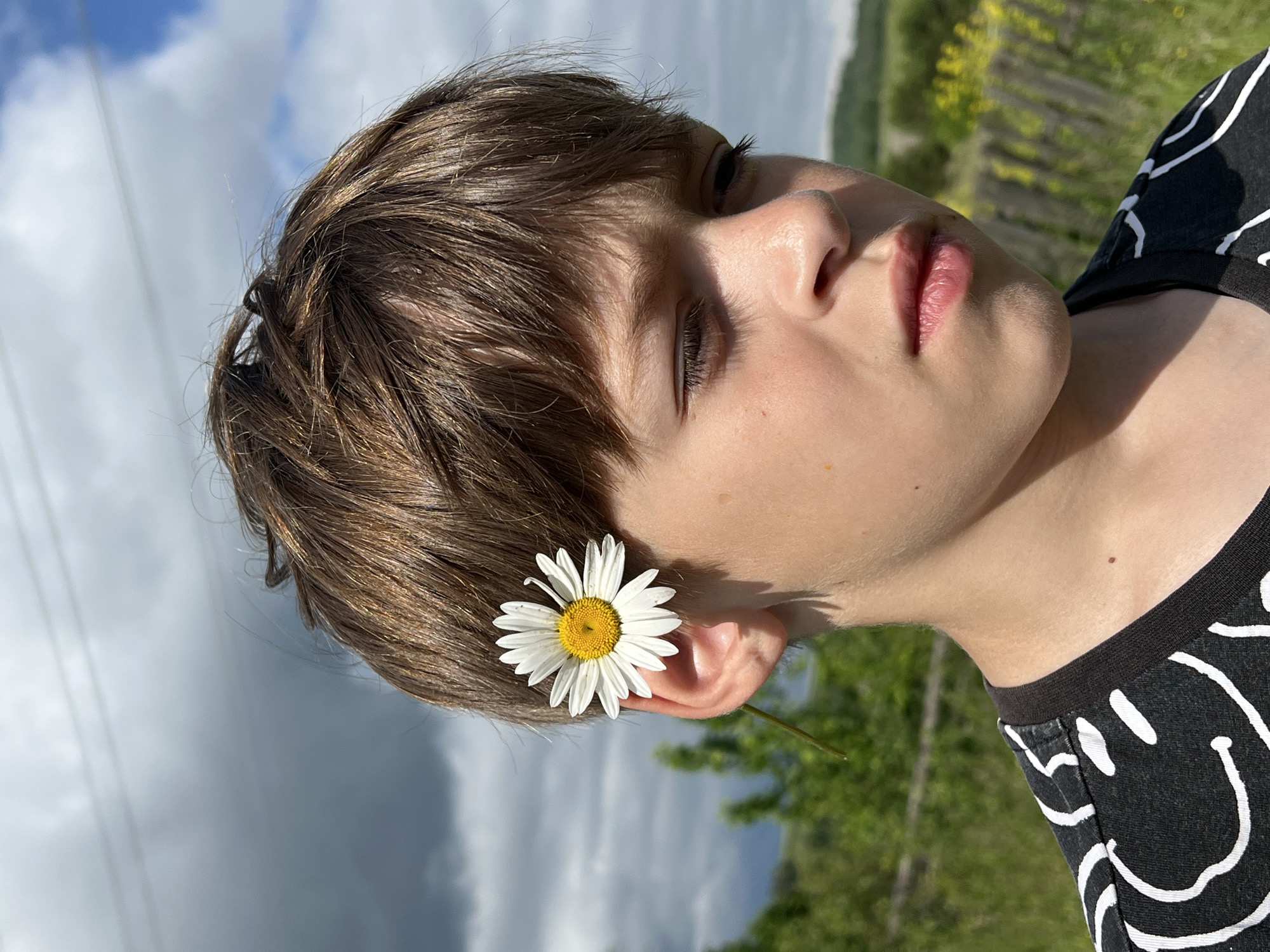 In an outdoor, natural setting, likely in Panteleevka, Russia, a young person with light brown hair is shown in a close-up portrait. A white daisy with a yellow center is tucked behind their right ear. The subject is wearing a black t-shirt with a white, abstract smiley face pattern. Their gaze is directed downwards and to the right. The background consists of green foliage and a fence, with a cloudy sky overhead.