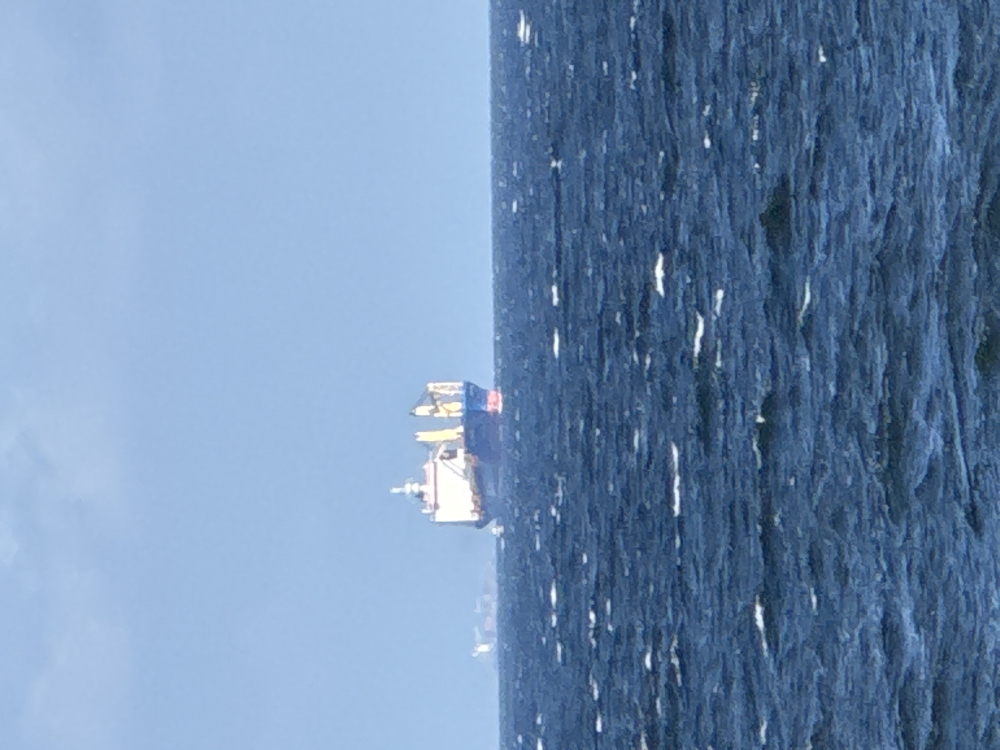 A cargo ship is visible in the distance on a choppy, deep blue ocean. The ship is white and blue with red accents on its hull. The sky above is a clear, light blue. The image appears to have been taken from a beach or shoreline. The ship is too far away to determine the specific type of cargo it is carrying. The lighting suggests it is daytime, likely overcast or with the sun behind the viewer. There are no people visible in the image, and no interaction is occurring. The environment is purely oceanic with a clear sky.