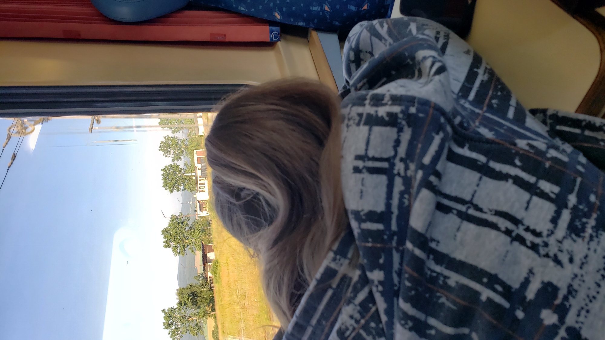 A person with long, blonde hair is resting their head on their arm and shoulder, wrapped in a blue and white patterned blanket. They are looking out of a train window at a rural landscape under a clear blue sky. The landscape features dry, yellowish grass, several green trees, and a few small, light-colored buildings, likely in Vydrník, Slovakia. The train car's interior is visible, with a yellow wall and a glimpse of a blue patterned seat or fabric. The lighting suggests it is daytime, possibly late afternoon.