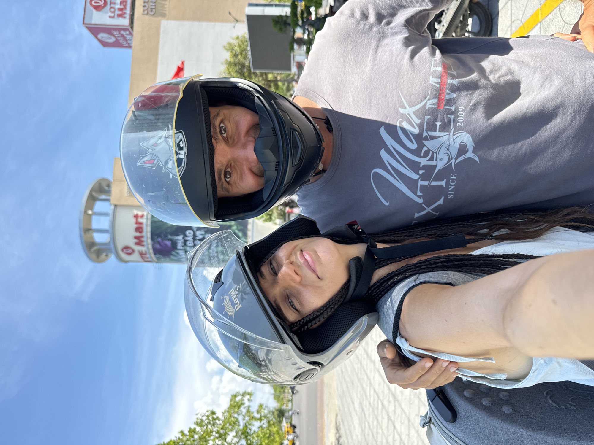 A man and woman, both wearing motorcycle helmets, are posing for a selfie outdoors. The woman is on the left, with her head tilted slightly and a faint smile. Her hair is in long braids. The man is on the right, looking directly at the camera with a broad smile. He is wearing a grey t-shirt with a "Max Extreme Since 2009" graphic. In the background, a Lotte Mart building and a billboard are visible, suggesting the location is within a commercial area of Phu Thuy Ward, Vietnam. The sky is clear and blue with some clouds.