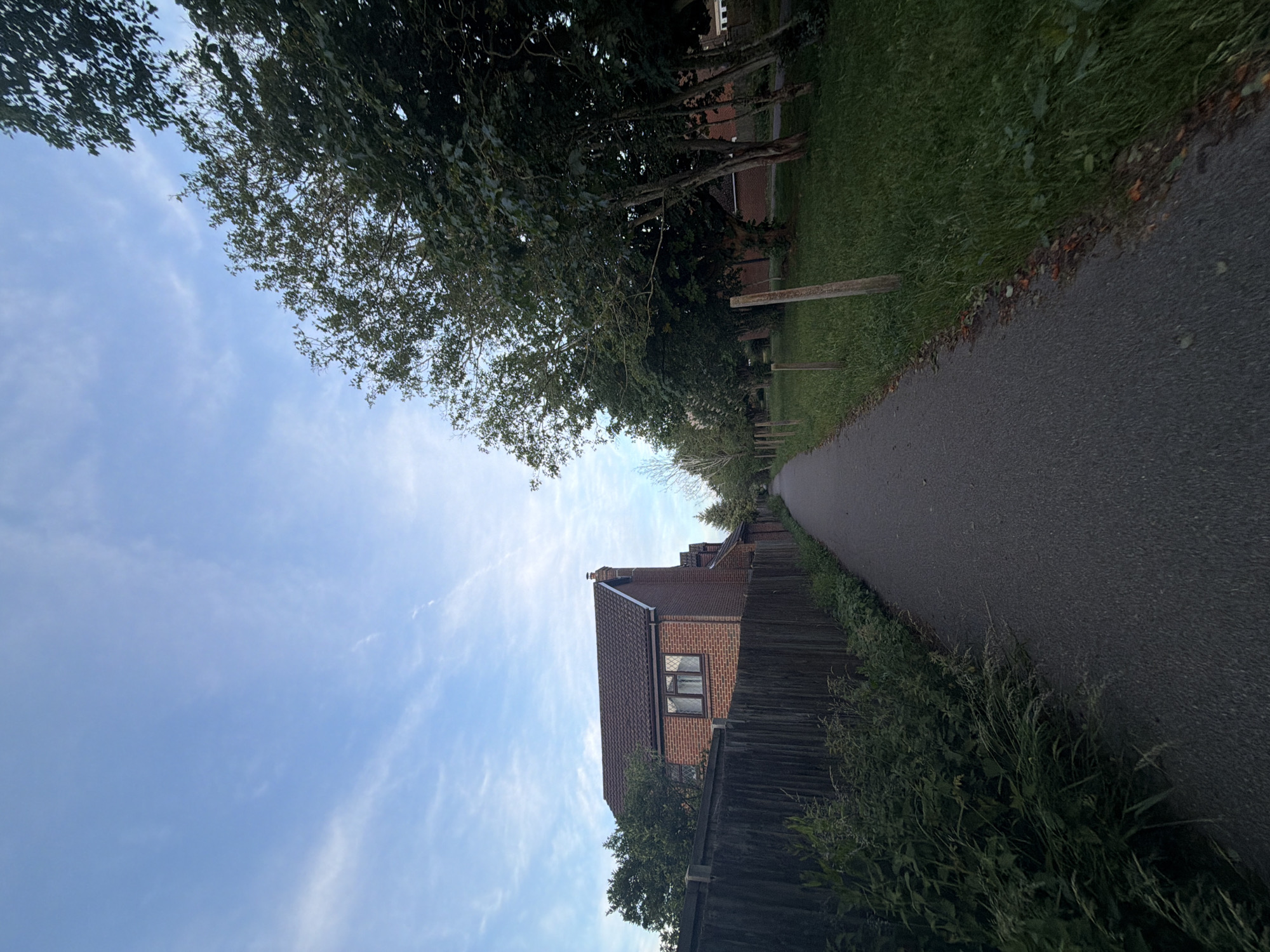 The image displays a straight, asphalt footpath or cycle path extending into the distance under a light blue sky dotted with scattered thin white clouds. This scene is located in a suburban residential area of Maidstone, United Kingdom.
On the left side of the path, a dark, weathered wooden fence runs parallel to the path. Behind this fence, a red-brick house with a dark tiled roof is partially visible, featuring a prominent brick chimney on its right side and a window with leaded glass and internal curtains. Dense green foliage, including overgrown weeds and shrubs, lines the base of the fence and slightly encroaches onto the path.
On the right side of the path, a strip of well-maintained green grass is separated from the path by a series of evenly spaced, short wooden posts. Beyond the grass, a line of mature deciduous trees with full green leaves stands, partially obscuring further brick houses in the distance. The lighting suggests either early morning or late afternoon, with soft shadows and illumination. No people or animals are visible, indicating a quiet environment.