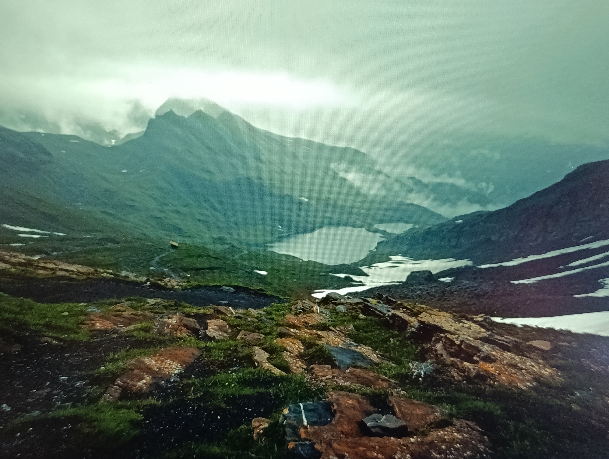 The image depicts a mountainous landscape under a cloudy, overcast sky. In the foreground, there is a rocky, uneven terrain with patches of green grass. Further into the scene, rolling green hills lead to a valley containing two bodies of water, likely lakes, with patches of snow or ice visible on the surrounding slopes. Distant, imposing mountains rise in the background, shrouded in mist and clouds. The overall atmosphere is cool and somewhat dramatic, suggestive of a high-altitude environment. There are no people or man-made structures prominently visible. The image is not from the city of Dubăsari town council, Moldova, as the scenery is characteristic of alpine or mountainous regions, not the topography of Moldova.