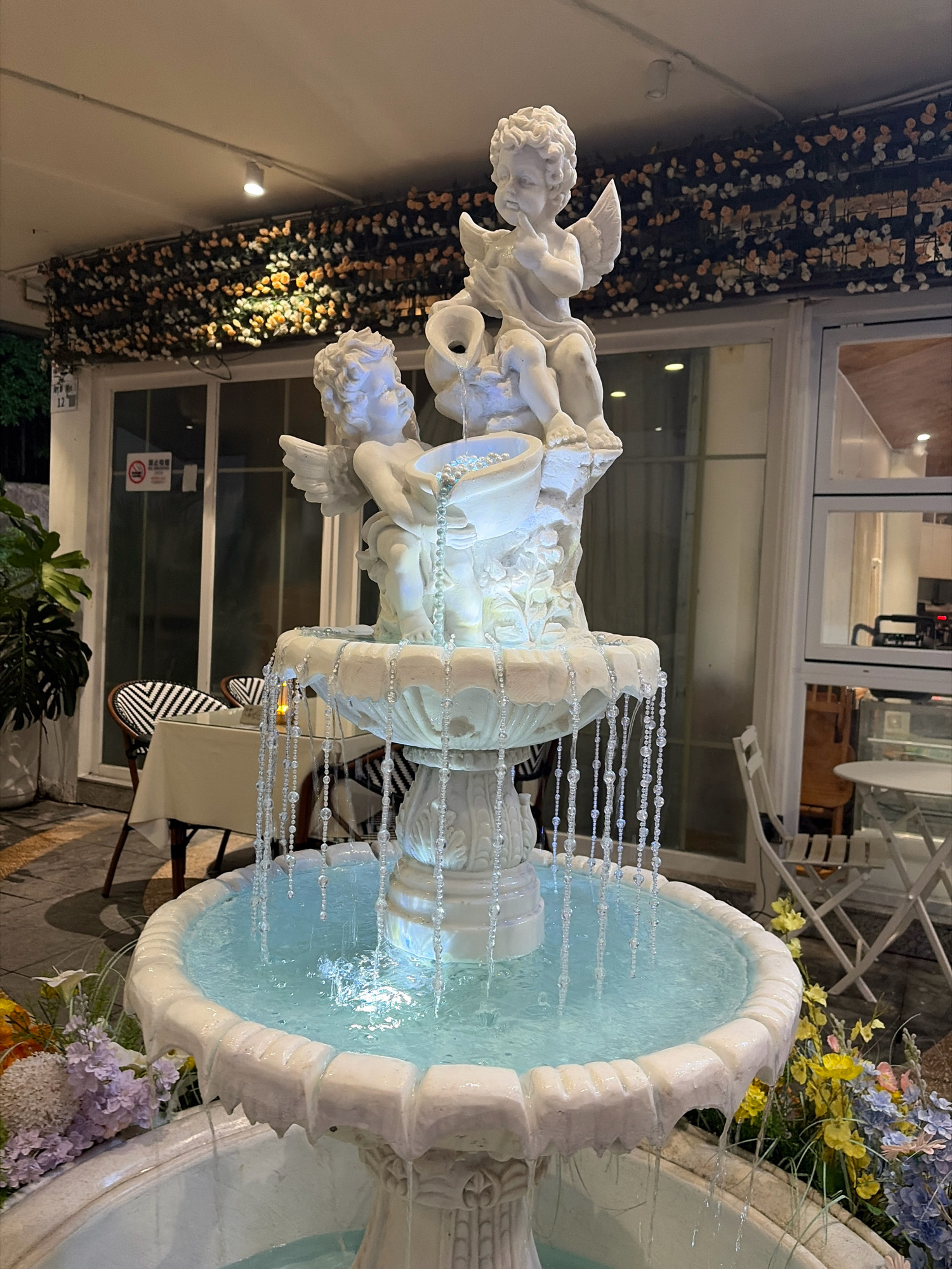 This image depicts a decorative fountain with cherub statues, likely located in a cafe or restaurant in Yantian District, China. The fountain is the central subject, featuring two white cherub figurines seated on a tiered structure, with water flowing from their pitchers. The fountain is illuminated and adorned with hanging beaded strands. Surrounding the fountain, we see tables and chairs set up outdoors, suggesting an dining or seating area. In the background, a building with large windows and an overhang decorated with artificial flowers is visible. Plants, including a large potted plant and floral arrangements, are placed around the fountain and seating area. The lighting suggests it is evening or a dimly lit indoor space.