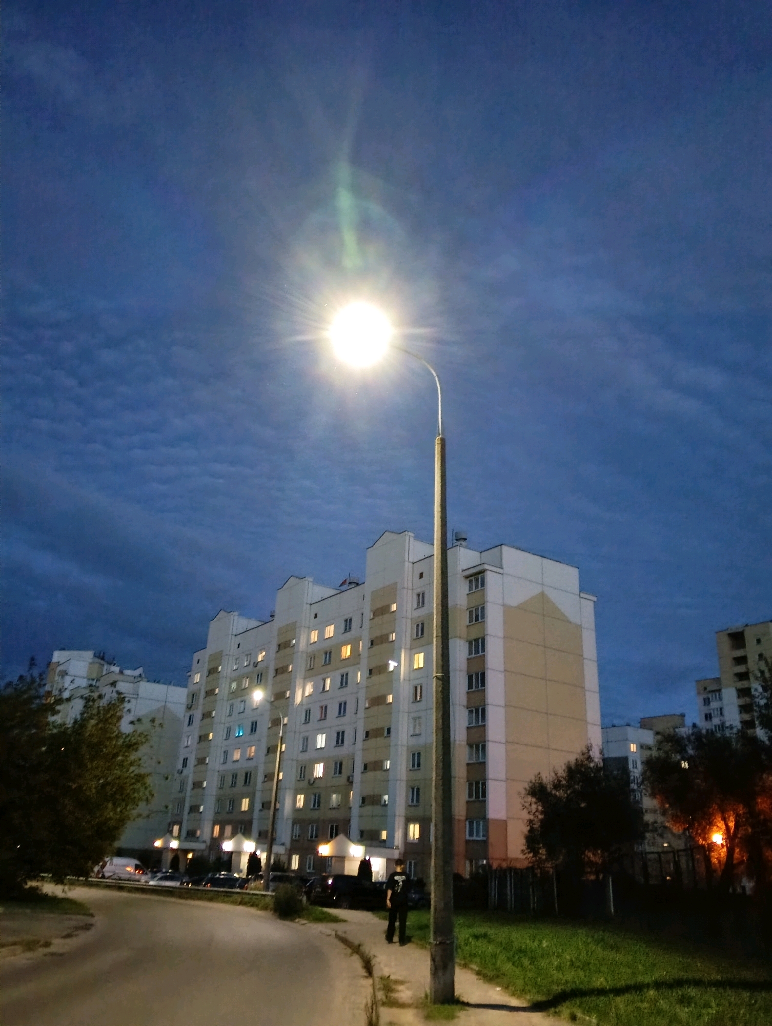 A low-angle, vertical, outdoor, nighttime photograph captures a street scene in a residential area of Hrodna, Belarus. A bright, circular street lamp, positioned in the center-right of the frame, dominates the upper portion of the image, emitting a strong glare that creates lens flare and washes out details in the sky. The sky above is a deep blue with faint, textured cloud formations and a greenish glow emanating from the lamp.
In the mid-ground, a multi-story, pale yellow and white apartment building stretches horizontally across the frame, its numerous windows illuminated from within. To the left of this building, another apartment building of similar design is partially visible. In the foreground, a paved road curves from the left into the scene, with a sidewalk and a grassy area on the right. Several parked cars are visible on the road in front of the apartment buildings.
A lone individual, dressed in dark clothing, stands on the sidewalk on the right side of the road, facing away from the camera. To the right of the sidewalk, trees with orange-tinted foliage are illuminated by an unseen light source. The overall impression is one of a quiet, residential evening in an urban setting.