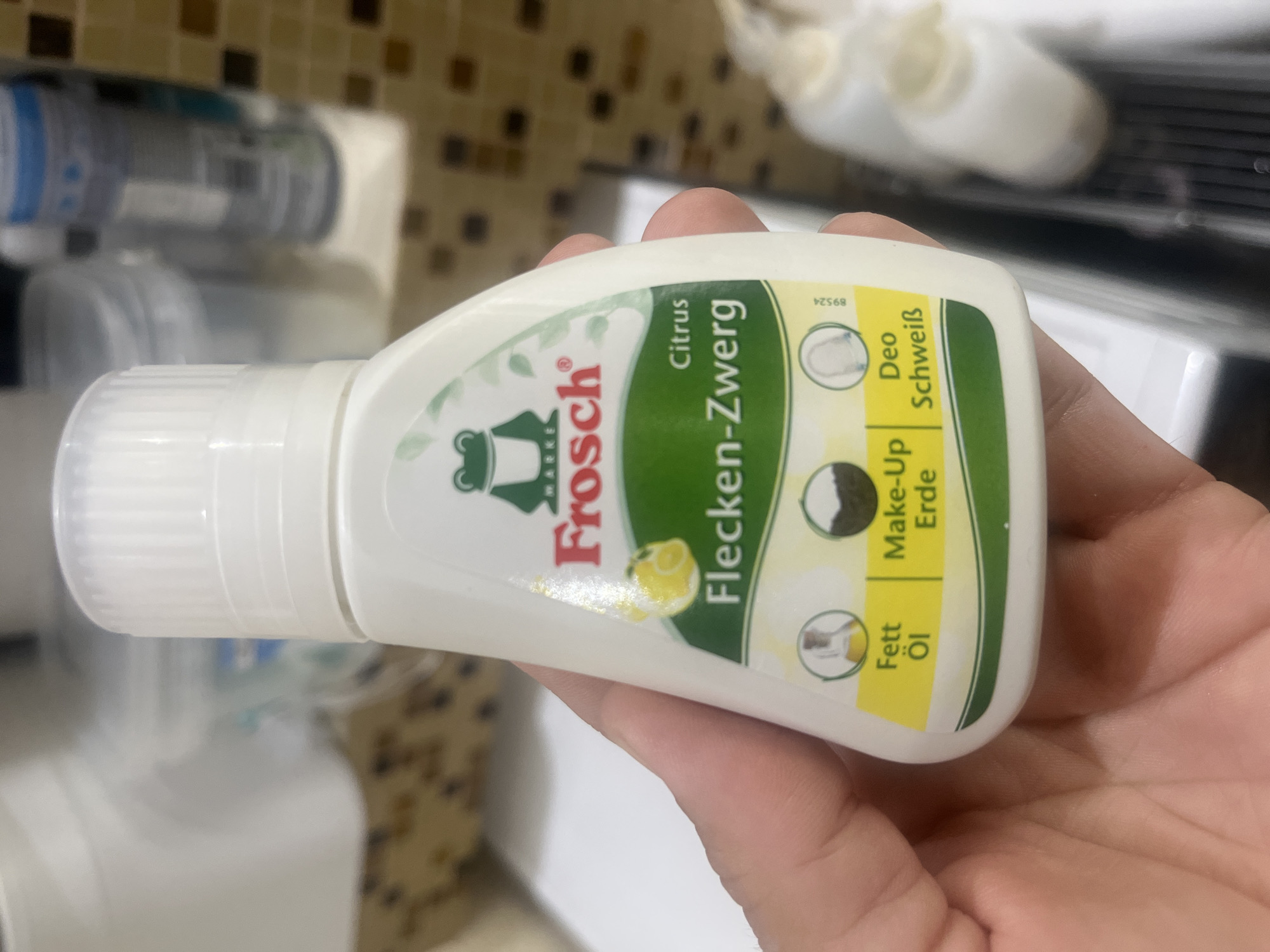 The image shows a person's hand holding a bottle of Frosch Flecken-Zwerg stain remover with a citrus scent. The bottle is white with a green label featuring a frog logo and text indicating it's suitable for grease, make-up, and deodorant stains. The background is blurred and appears to be a bathroom shelf with other toiletries. There is no information in the image that indicates its location in Onokivtsi, Ukraine.