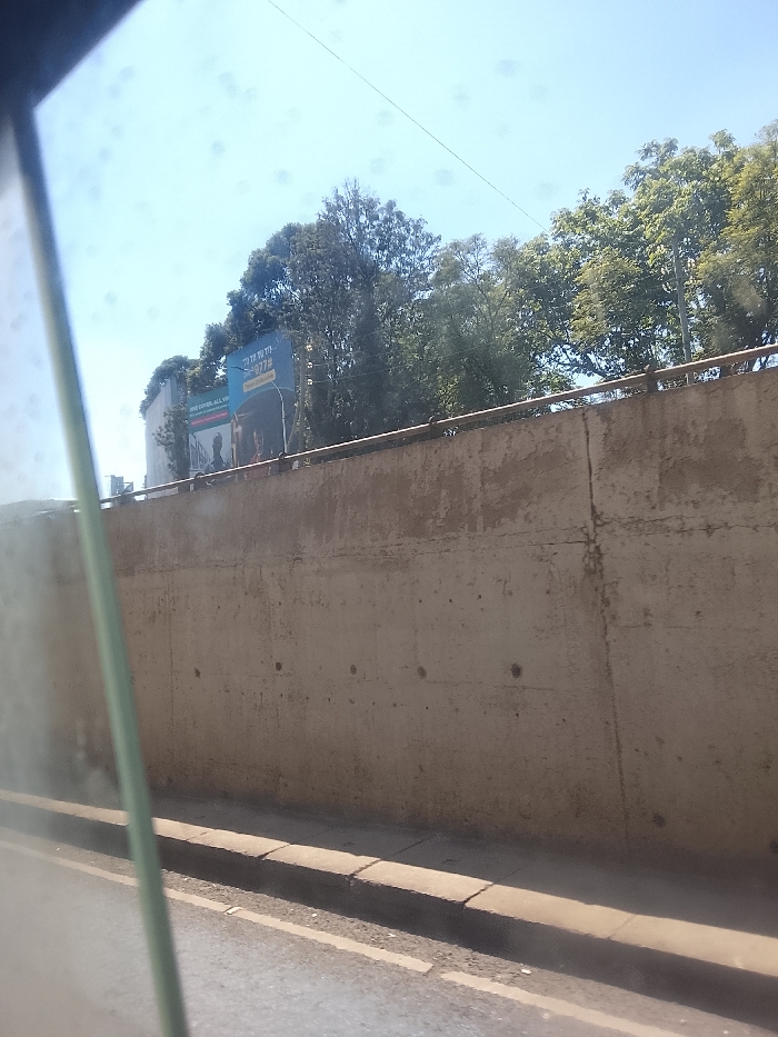 The image, captured from a moving vehicle, presents a roadside scene under a clear blue sky. In the immediate foreground on the left, a blurred, vertically oriented green or gray element partially obscures the view, suggesting a car window frame or pillar. To its right, a section of asphalt road is visible, marked by faint white lines. Adjacent to the road is a concrete curb, leading to a textured, light brown concrete retaining wall that dominates the midground. The wall appears aged and has visible seams and minor irregularities. A simple metal railing runs along the top of this wall.
Above and behind the wall, a dense canopy of green trees stretches across the upper part of the image, with some lighter-colored foliage towards the right, indicating bright sunlight. Nestled among the trees and partially visible above the wall on the left side of the background is a large billboard. The billboard features a blue section at the top with white text that reads "IT'S UP TO YOU" and a number "9772" below it. Further down on the billboard, on a darker background, more text is partially legible, appearing to say "ONE COVID, ALL V..." or "STOP COVID, ALL V...". A silhouette of a person is also visible on this lower section of the billboard, suggesting a public awareness campaign.
The bright, unfiltered sunlight suggests it is daytime, likely mid-morning or early afternoon, with no clouds in the sky. No people are explicitly visible in the scene, only the stylized figure on the billboard. The overall impression is an urban or suburban setting, possibly in Kiambu Township ward, Kenya.