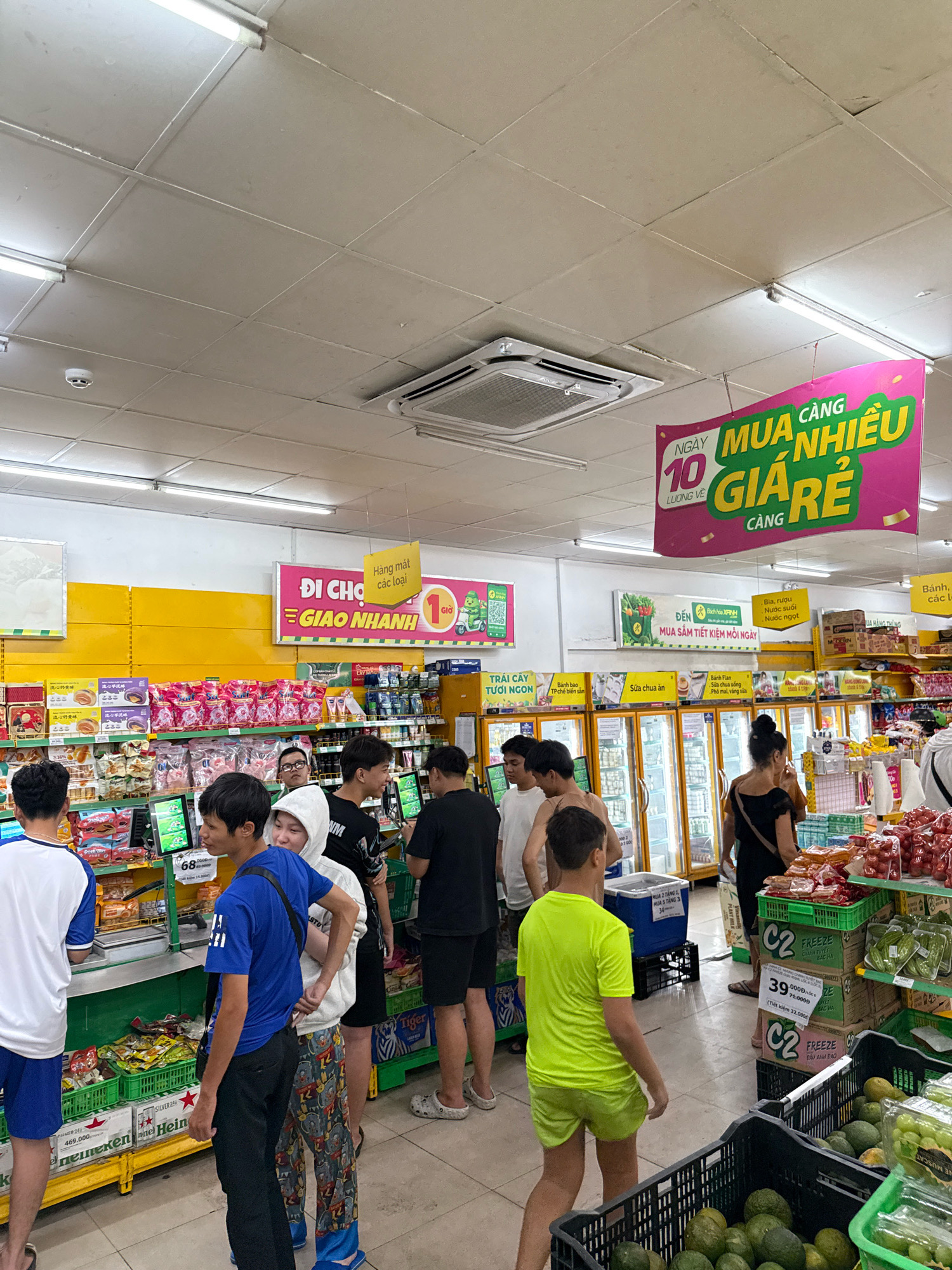 This is a medium shot, taken from a low angle, inside a brightly lit grocery store. A group of young men and a woman are browsing the aisles, looking at various products. In the background, there are rows of refrigerators stocked with drinks and a large display of fresh produce. A promotional banner hangs from the ceiling, advertising a sale with the text "NGAY 10 CANG MUA NHIEU GIA RE". The floor is tiled, and the ceiling has fluorescent lighting. The overall atmosphere appears to be casual and busy, typical of a local market. The presence of Vietnamese text on signage and product packaging indicates the location is likely in Vietnam. Given the context and typical shopping habits, this scene could plausibly be situated in Nha Trang, Vietnam, a popular coastal city with numerous supermarkets and markets.