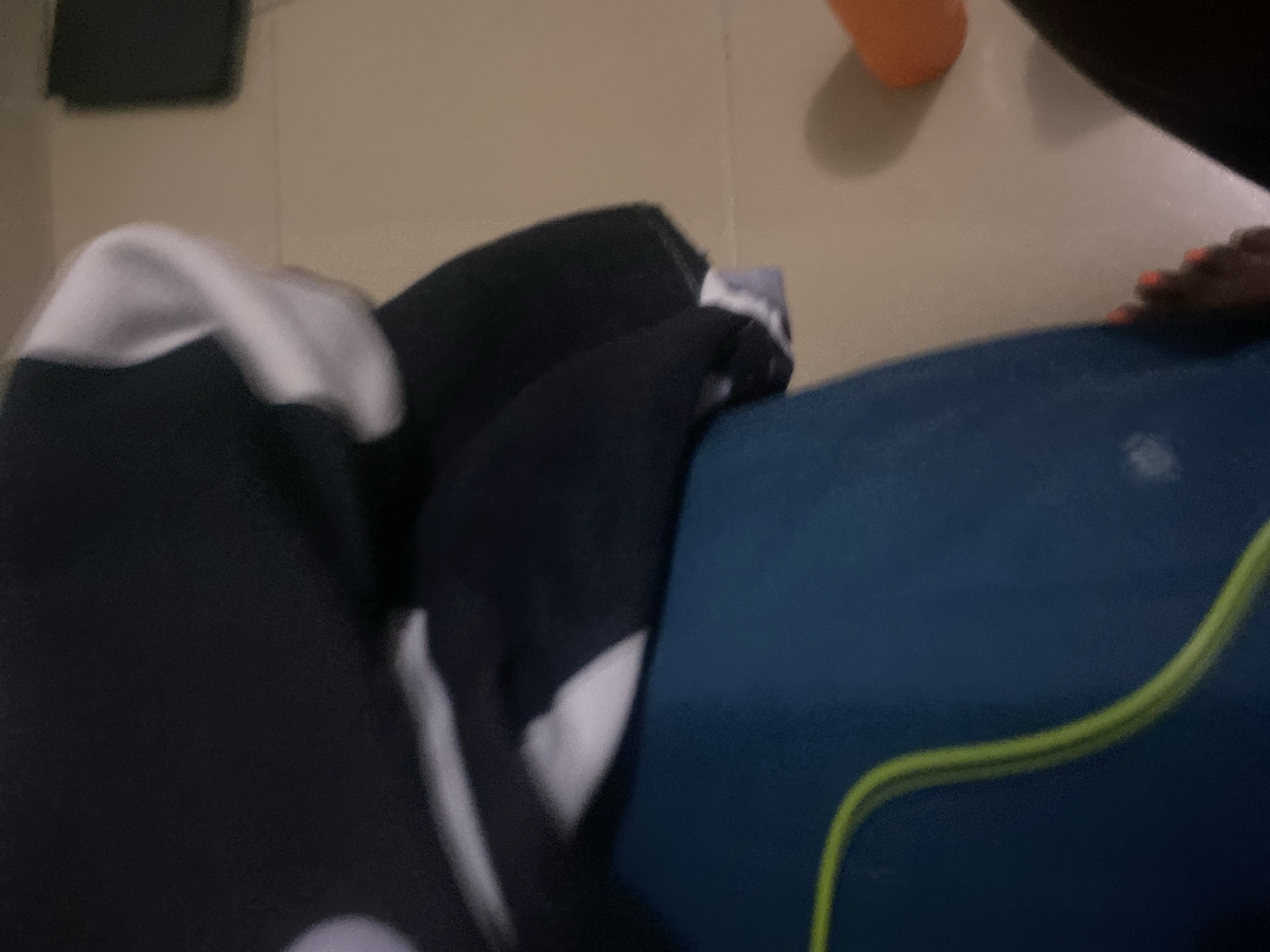The image shows a close-up view of a person's feet and legs wearing blue athletic shorts with a neon green stripe. They are standing on a tiled floor, possibly in a bathroom or shower, with a towel draped over their legs. An orange object is visible in the background, possibly a bath product. The lighting is dim. There is no visible indication of the location being Howayi, Nigeria.