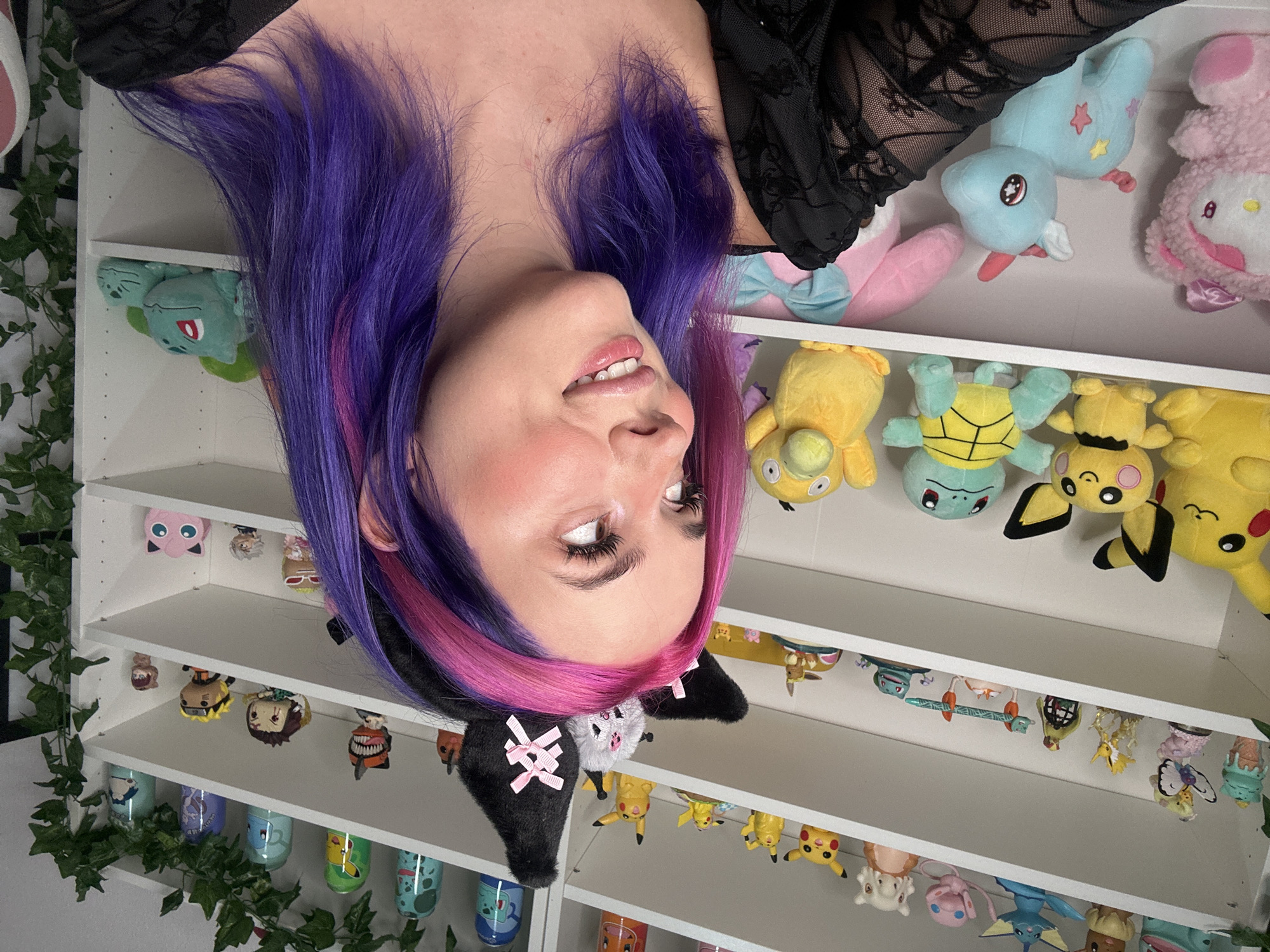 This is a medium shot, eye-level, interior photograph taken in Oviedo, Spain. The image features a young woman with brightly colored pink and purple hair, wearing black lace clothing and a black headband with ears adorned with pink bows. She is looking to her right with a slight smile. Behind her, a white shelving unit is filled with various collectible figurines and plush toys, primarily characters from Pokémon and other anime series. The shelves are organized and well-lit. The environment appears to be a room dedicated to displaying these collectibles. The overall tone is bright and cheerful.