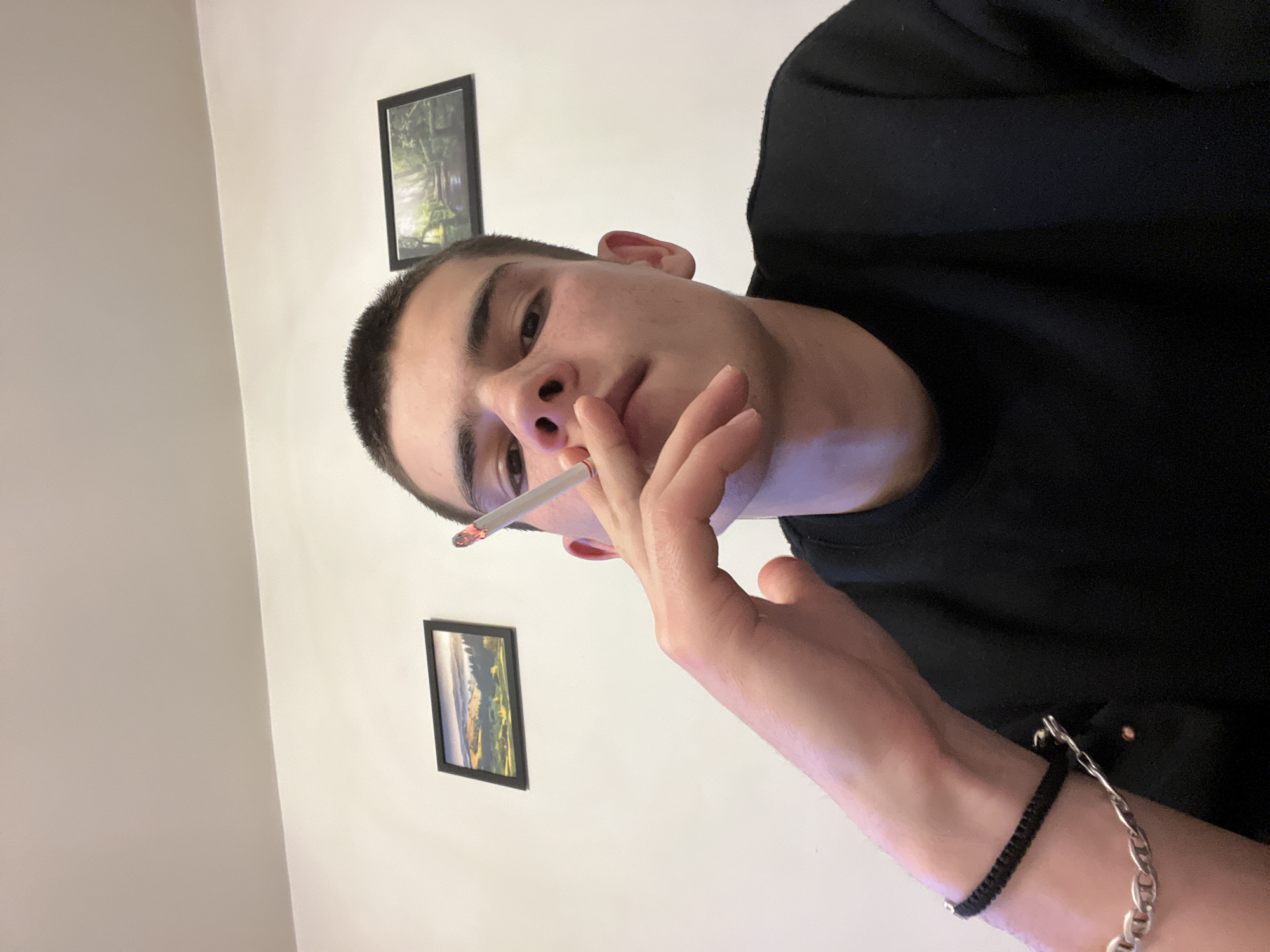 A young man with short dark hair is holding a lit cigarette to his mouth. He is wearing a black shirt and a black and silver bracelet on his left wrist. Behind him are two framed pictures on a white wall. There are no indications of the image being related to the city of Plovdiv, Bulgaria.