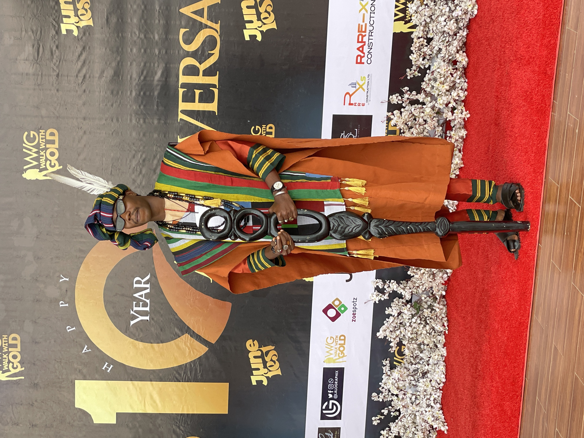 A man stands on a red carpet in front of a black backdrop at the "Walk With Gold" event in Barakin Lerawi, Nigeria. The man is wearing traditional Nigerian attire, including an orange robe with colorful striped trim, a striped headwrap adorned with white feathers, sunglasses, and beaded necklaces. He holds a carved wooden staff with circular elements. The backdrop features the event logo, "Walk With Gold," and the text "Happy 10 Year Anniversary." The carpet is lined with artificial white flowers. Several sponsor logos are visible on the backdrop and to the left of the man. The floor appears to be made of wooden planks.