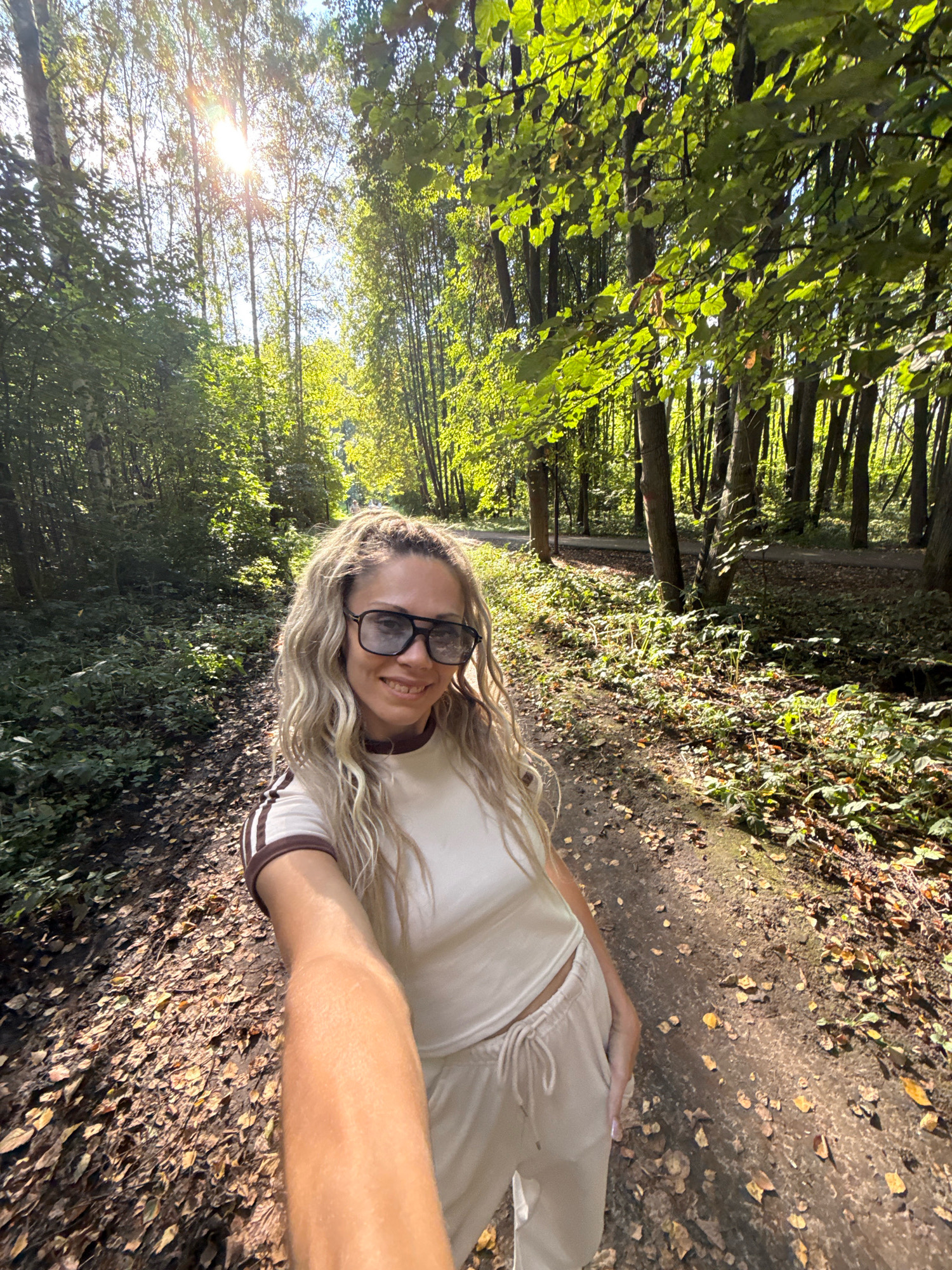 A woman with blonde, wavy hair and sunglasses is taking a selfie while walking on a path in a forest. She is wearing a light-colored t-shirt with brown stripes on the sleeves and matching light-colored sweatpants. The path is covered in fallen leaves, and the surrounding area is filled with trees and greenery, illuminated by sunlight filtering through the canopy. The scene appears to be in Rusanovo, Russia.