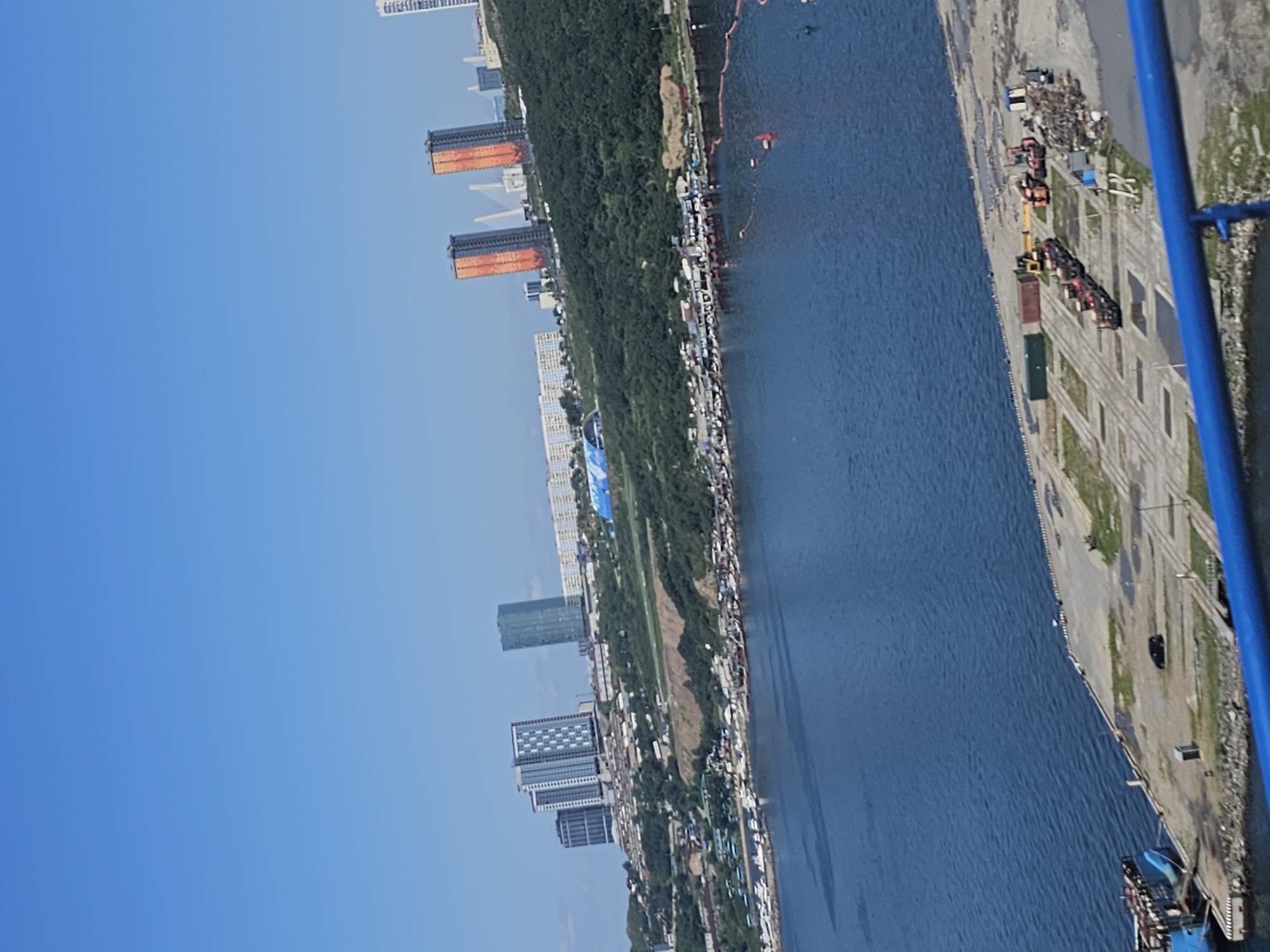 This is an aerial view of a waterfront in Vladivostok, Russia, under a clear blue sky. The foreground shows a blue railing, indicating the viewpoint is likely from a balcony or elevated platform. To the left, a portion of a blue ship is docked at a concrete pier. The pier itself is sparsely populated with a dark-colored vehicle, some seagulls, and a small utility building. Further into the frame, industrial equipment, including a yellow excavator, red cranes, and shipping containers, are visible on the pier.
The middle ground depicts a bustling waterfront area with numerous small boats and vessels moored along a shoreline dotted with buildings. Beyond this, a densely forested hillside rises, leading to a cityscape. Prominent in the cityscape are several tall buildings, including a cluster of modern high-rises and two striking orange-hued skyscrapers that are characteristic of Vladivostok's skyline, likely near the Golden Horn Bay. In the distance, a large suspension bridge with white towers is visible, a notable landmark in the city. The overall scene captures a mix of industrial activity, urban development, and the natural landscape of Vladivostok.