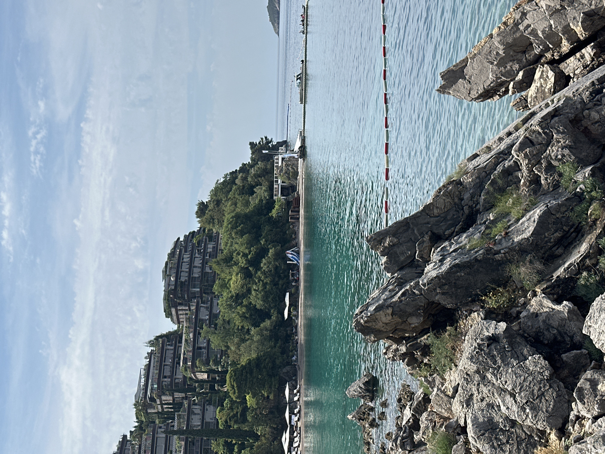 A wide shot captures a scenic view of a bay in Budva, Montenegro. In the foreground, weathered grey rocks jut out into the turquoise water. A red and white buoy line floats on the surface, marking a boundary. The middle ground features a sandy beach with rows of white lounge chairs and umbrellas, suggesting a resort or private beach area. A set of blue and white water slides is visible on the beach. Lush green trees and vegetation cover the hillside that rises behind the beach. Dominating the upper portion of the hillside are several multi-tiered, modern buildings with balconies and dark-tinted windows, integrated into the natural landscape. The buildings appear to be a hotel or resort complex. In the distance, the calm sea stretches towards the horizon, with a small island visible on the right. The sky is partly cloudy with a soft blue hue.
