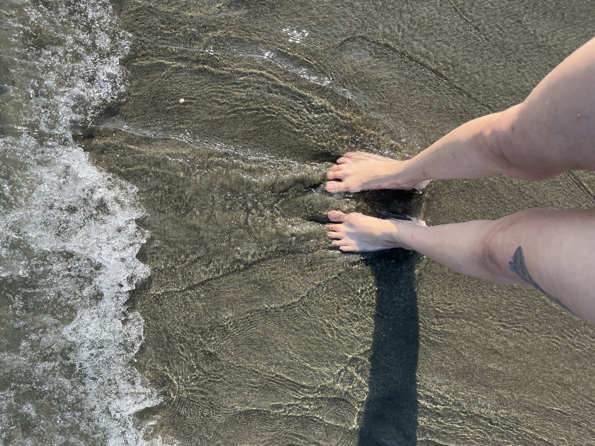 The image shows a person's bare feet and legs standing at the edge of the sea on a sandy beach. The water is shallow and clear, with small waves breaking over the sand. The sunlight creates reflections and ripples on the water's surface. The sand appears dark and wet, with visible patterns from the receding water. A shadow of the person's legs is cast on the wet sand to the left. A tattoo of a stylized feather or wing is visible on the person's left thigh. The location is identified as Agios Tychona Community, Cyprus.