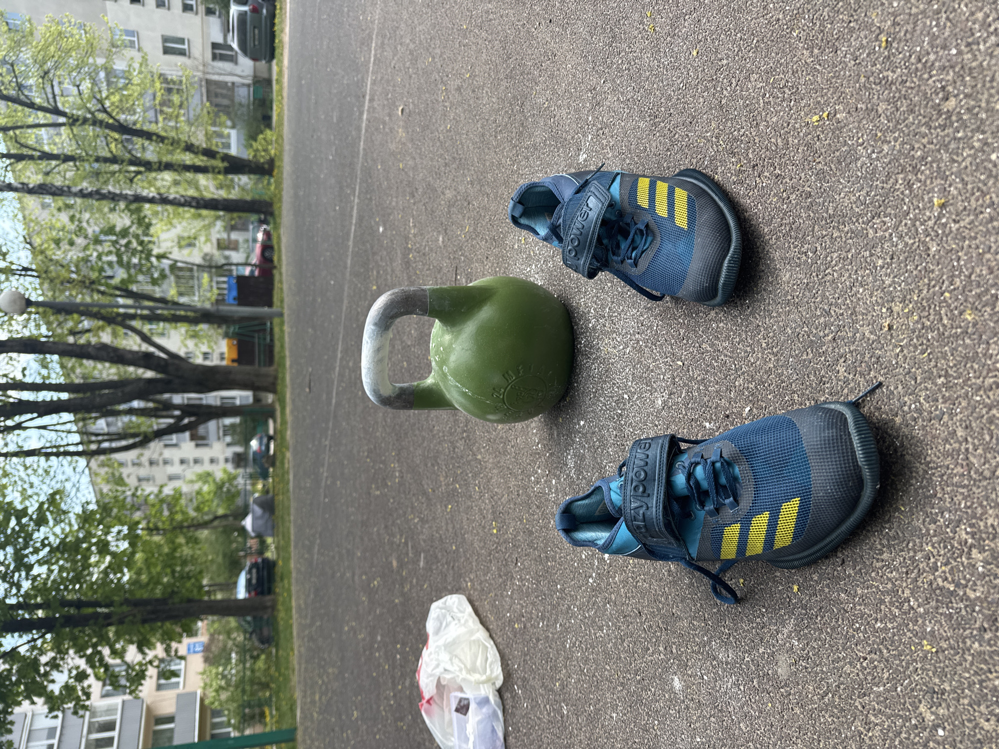 A kettlebell and a pair of weightlifting shoes sit on a paved surface. The kettlebell is olive green with a silver handle. The shoes are blue with yellow stripes and black straps labeled "ADZYPOWE". A translucent plastic bag lies on the lower left. In the background, trees and residential buildings are visible, suggestive of an outdoor exercise area in a residential area. The location is Minsk, Belarus.