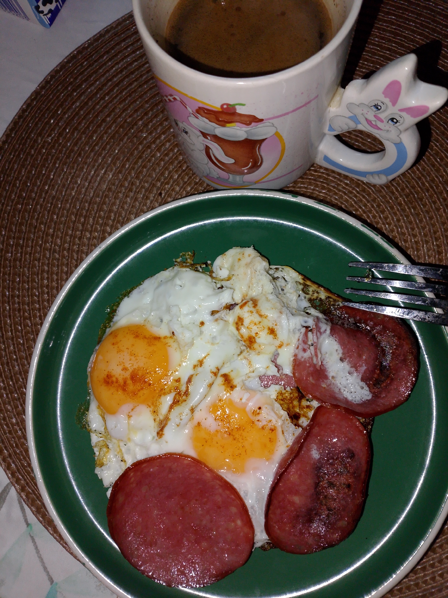 A plate of fried eggs and sausage sits on a brown woven placemat. A mug of coffee is placed above the plate. A fork lies partially on the eggs and sausage. A corner of what appears to be a milk carton is visible in the upper left corner. The plate is green with a white border. The mug is white with a cartoon design and a rabbit-shaped handle. The scene is well-lit. No location details in Izhevsk, Russia, are detectable in this photo.