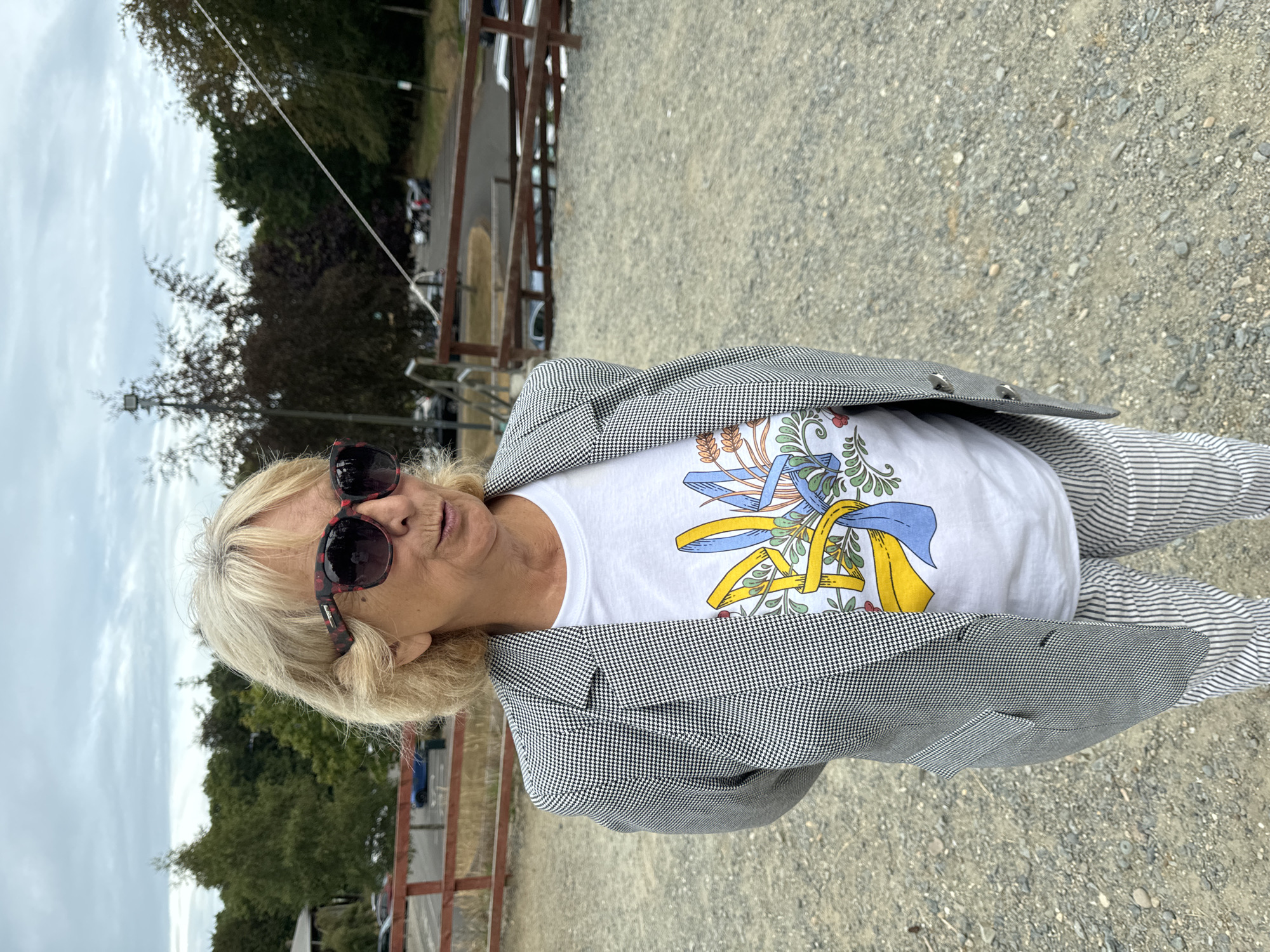 A medium shot shows a woman from the chest up, standing outdoors on a gravel surface. She has blonde hair and is wearing large sunglasses with a red and black patterned frame. She is dressed in a white t-shirt with a graphic design featuring a yellow and blue emblem, and a black and white houndstooth blazer. Her hands are clasped behind her back. Behind her, a wooden fence runs horizontally. Beyond the fence, trees line the background, and a cloudy sky is visible. The location is identified as Courtown, Ireland.