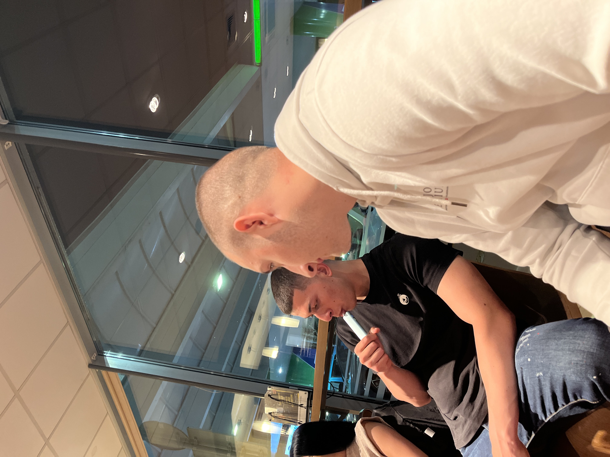 The image captures three individuals indoors. The main subjects are two men, one in the foreground with a shaved head and a white hooded sweatshirt, positioned so that most of his body is out of frame. The second man is wearing a black t-shirt and jeans, holding a vape pen or similar device. He seems to be looking down and holding the vape to his mouth. Only part of a third person is visible, wearing a light-colored top. They are sitting at a table with large windows and a view that suggests an airport or similar facility. The scene might be in Burgas, Bulgaria, due to the background elements visible through the windows, such as signs and building architecture.