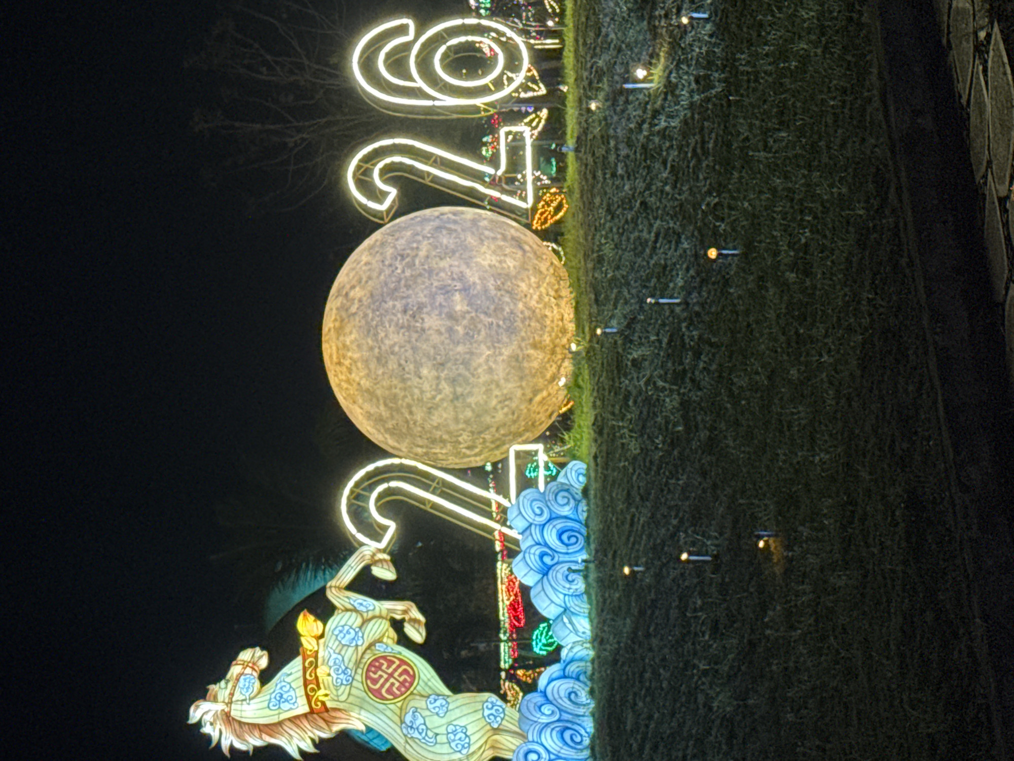 The image, taken at night, shows a display in what is likely Hội An Ward, Vietnam. A large illuminated sphere resembling a moon is centrally placed. Directly above and below the moon are illuminated numbers “2023,” indicating this image was taken during that year's celebratory display. Below the numbers is a horse figure, decorated with what appear to be Chinese-style art. To the right, running vertically, is grass with small lights along a path. The background is completely dark.