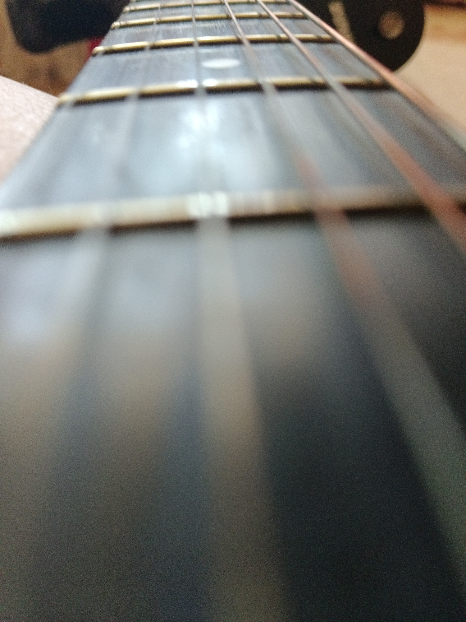 The image is a close-up, low-angle shot of a guitar neck. The strings are visible, running parallel to each other along the fretboard. The frets are also visible as thin, horizontal lines across the neck. The background is out of focus, showing a dark circular object and a blurry hand. The image appears to be taken in a dimly lit indoor setting. There are no visible indications of the city of Severnyi, Russia, or any specific environmental details that would suggest this location.
