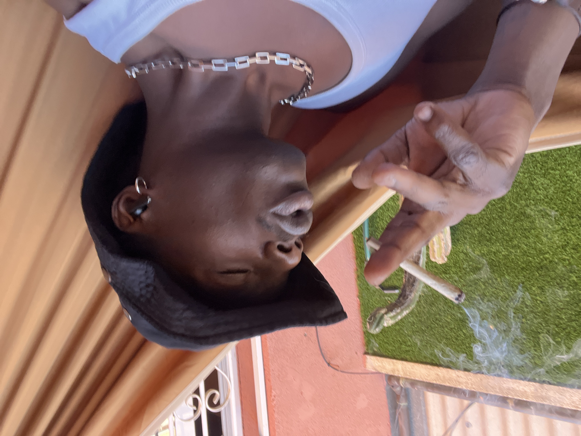 The image shows a person with dark skin, wearing a white tank top and a dark bucket hat. They are adorned with a silver chain necklace and an earring in their left ear, possibly accompanied by an earbud. Their eyes appear closed or mostly closed, suggesting a moment of relaxation.
The person's right hand is visible, holding a lit cigarette or joint from which smoke emanates and wafts downwards. Next to their hand, on a vibrant artificial green grass surface, a green lizard or similar reptile is prominently visible.
The setting appears to be an outdoor space, possibly a patio or balcony in Barcelona, Spain. The background features a reddish-brown wall, a light wooden ledge, and striped orange-brown fabric, which could be a curtain or awning, suggesting an area protected from direct sun. A white window frame with decorative metalwork is also visible in the lower left background. The lighting indicates it is daytime, likely sunny. No visible text is present in the image.