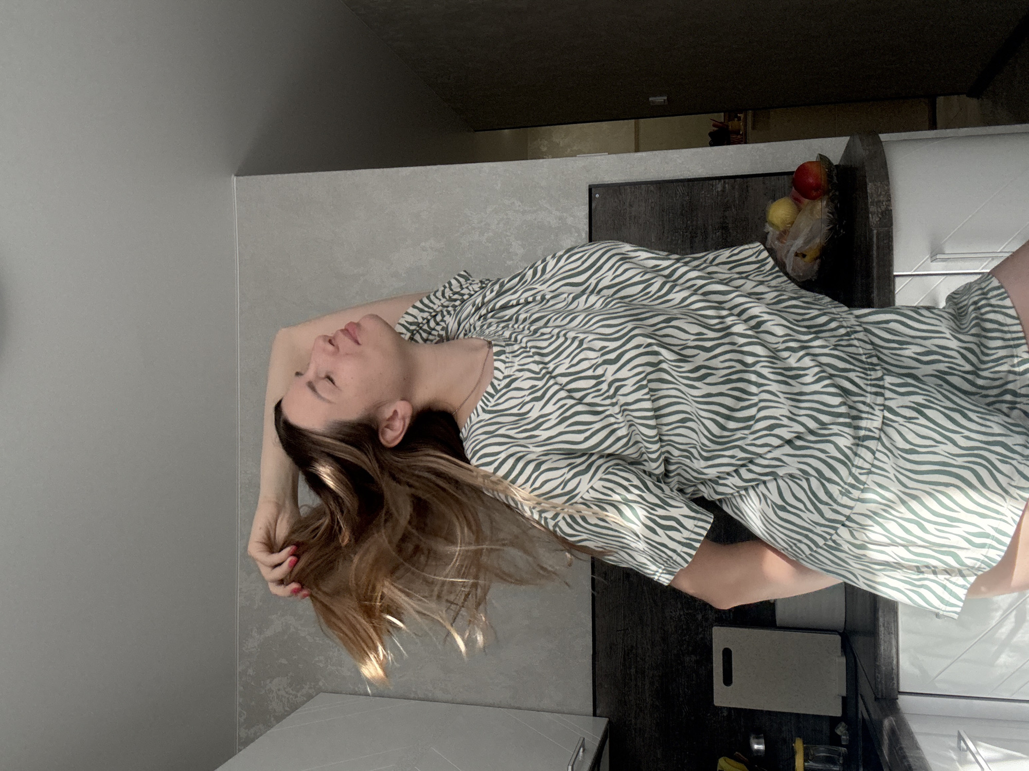 A woman with long, light brown hair is captured in a medium shot, standing in what appears to be a kitchen. She is wearing a pajama set with a green and white zebra-like print. Her right arm is raised behind her head, with her hand tangled in her hair, causing it to fan out. Her eyes are closed, and her head is tilted back slightly, suggesting a moment of relaxation or playful movement. The background shows kitchen cabinetry, countertops, and a fruit bowl containing apples and other fruits. The lighting suggests it is daytime, with sunlight streaming in from the right, creating some shadows and highlights on the woman and her surroundings. The overall setting is domestic and casual.
