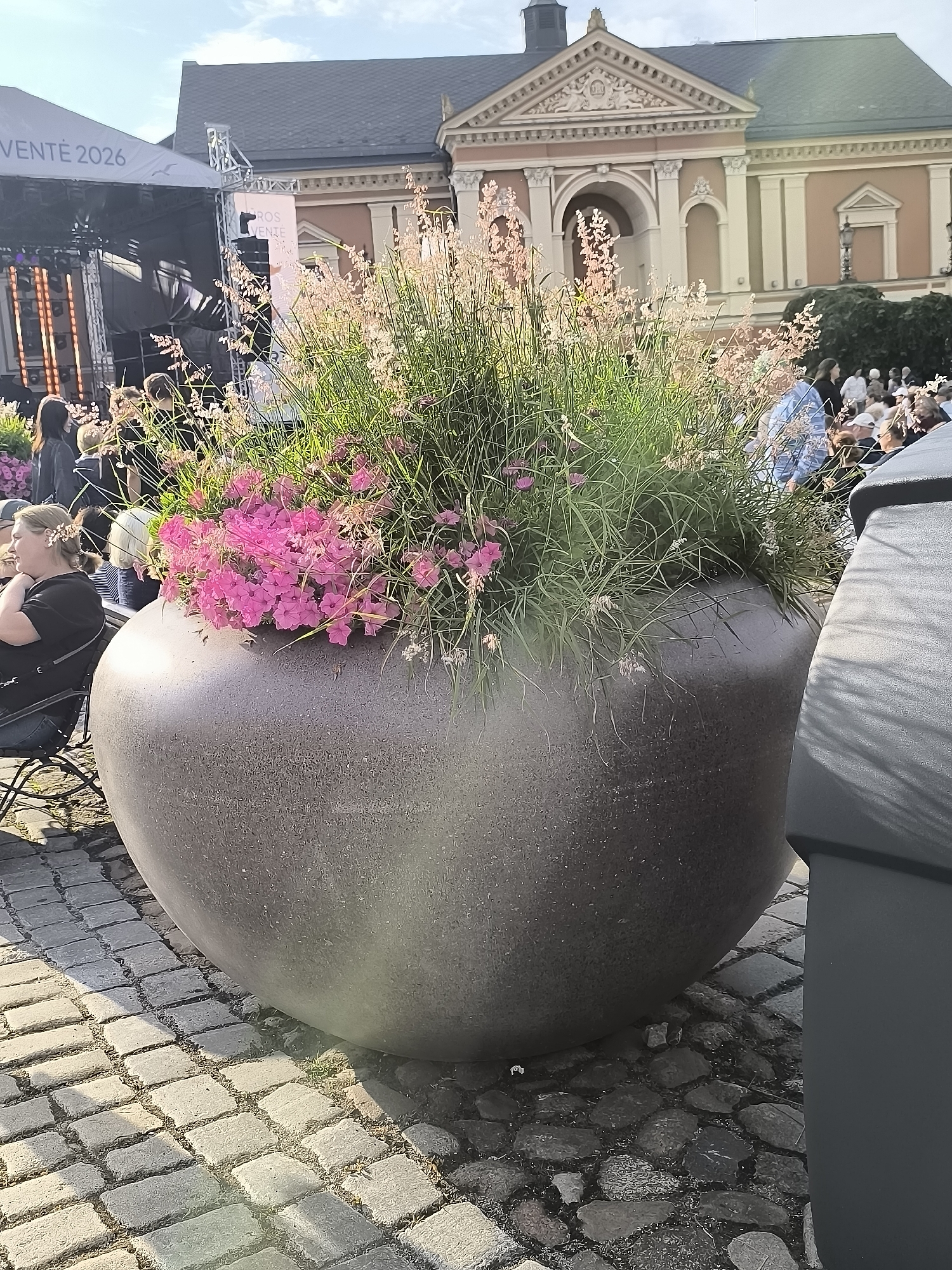 In the foreground, a large, dark, ovular planter overflows with green foliage and pink flowers. The planter sits on a cobblestone surface. To the left of the planter, a woman in a black shirt and jeans is seated, leaning forward. Behind her, a stage structure with a banner reading "VENTÉ 2026" is visible, along with various lights and equipment. In the background, a classical building with arched windows and ornate detailing stands under a bright sky. A crowd of people is gathered in front of the building, some sitting and some standing. To the right of the planter, a dark, rectangular object, possibly a trash receptacle, is partially visible. The scene appears to be an outdoor event in a public square in Klaipėda, Lithuania.