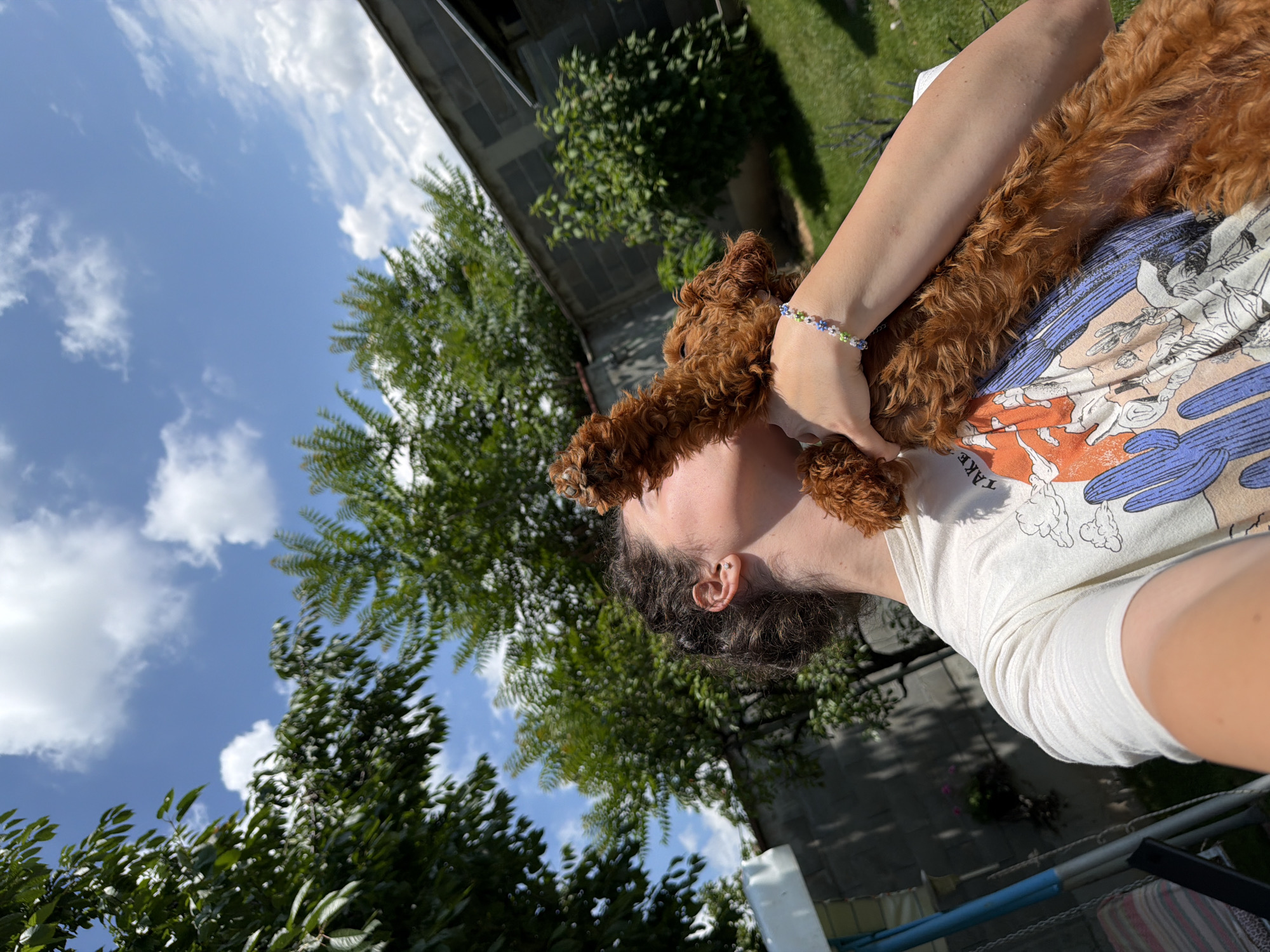 The media file is a low-angle, close-up, selfie-style shot of a person holding a small, fluffy, reddish-brown dog. The person is wearing a white t-shirt with a graphic print that includes clouds and a blue bird-like figure. A bracelet with small, colorful beads is visible on the person's wrist. The dog is nestled against the person's chest and neck, with its head near the person's face. The person's hair is dark and curly, and they have an earring in one ear.
The background is blurred but suggests an outdoor setting with lush green trees and a bright blue sky with fluffy white clouds. A brick building is partially visible in the upper left corner. The lighting indicates it is daytime, likely sunny. The overall mood appears relaxed and affectionate, with the person and dog seemingly enjoying a moment together in an outdoor environment. The provided location context is Sofia, Bulgaria.