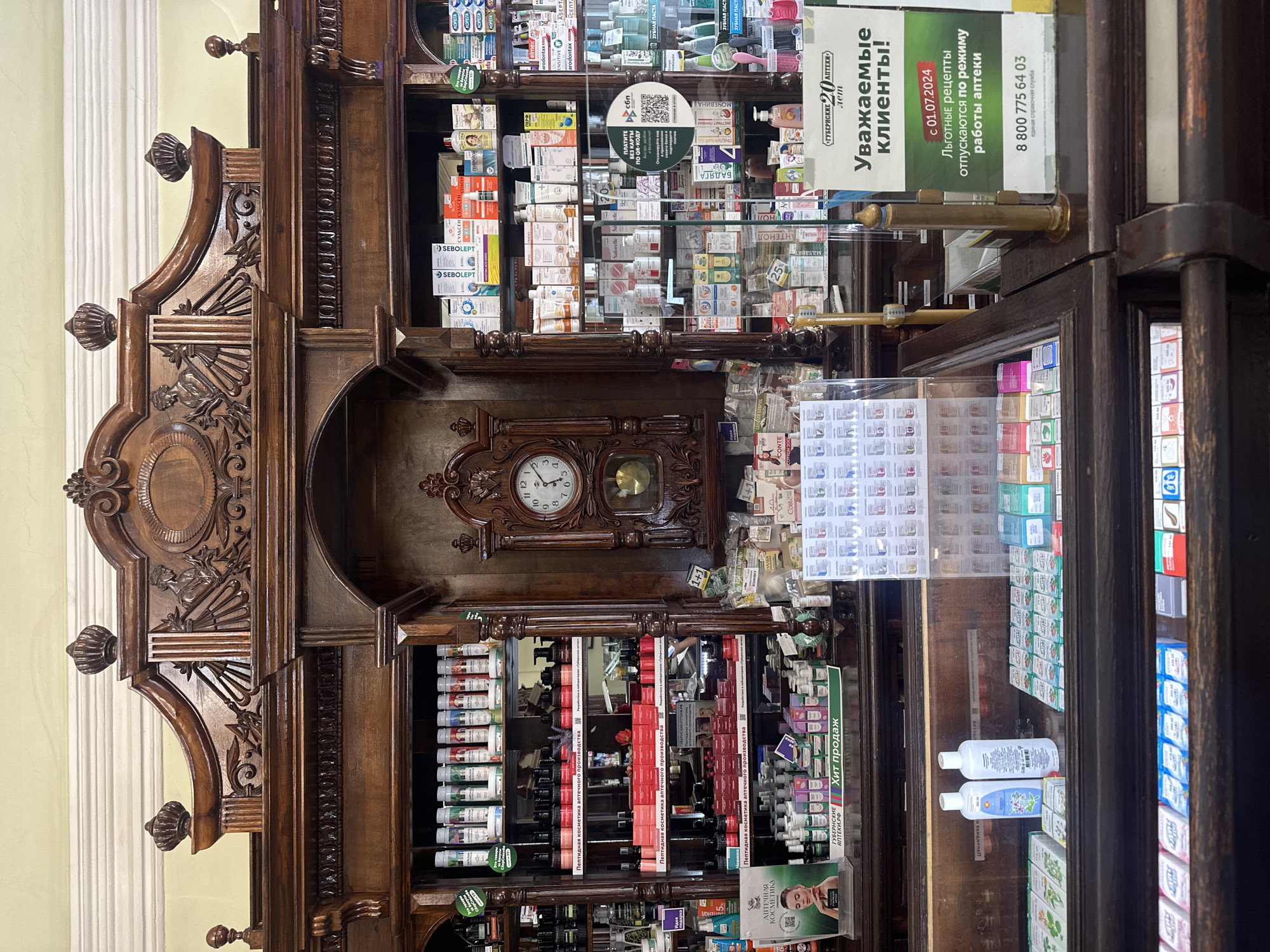 The image shows an interior view of the historical Gubernskiye Apteki (Губернские Аптеки) pharmacy in Krasnoyarsk, Russia. The foreground displays shelves stocked with various pharmaceutical and cosmetic products. The shelves are filled with boxes of medicines, tubes, and bottles, including items labeled "SEBOLEPT" and "Пептидная косметика аптечного производства" (Peptide cosmetics of pharmaceutical production). A sign that reads "Уважаемые клиенты!" (Dear customers!) and an announcement about prescription regulations from July 1, 2024, and a phone number is visible. An antique wooden display case dominates the background, featuring ornate carvings and a vintage clock. The setting suggests a blend of historical preservation and commercial activity.