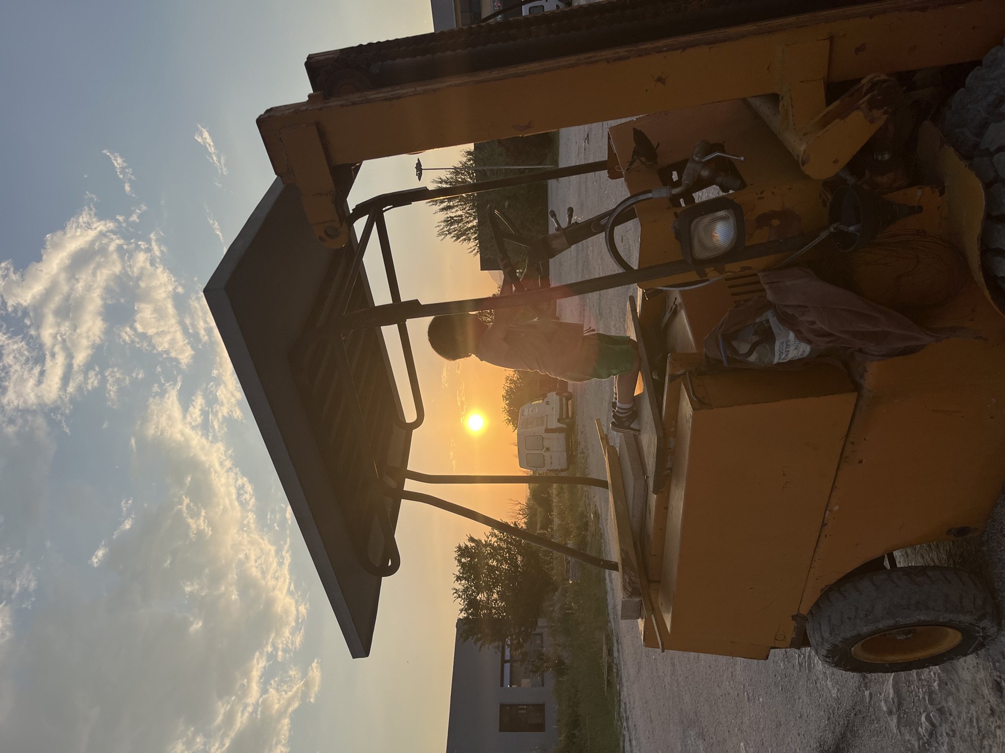 A child is standing on the seat of a yellow forklift, silhouetted against the setting sun. The sky is pale blue with white clouds on the left side of the frame. The forklift is parked on a gravel surface in Pobrdje, Montenegro. In the background, there are trees, a white van, and a building with a dark gray facade.