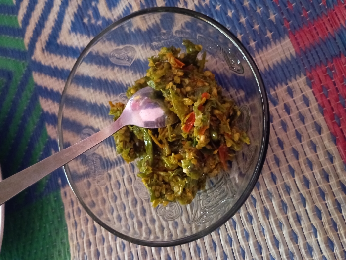 An overhead close-up shot features a clear glass bowl containing a green chili paste or relish, commonly known as sambal ijo, placed on a patterned woven mat. A silver metal spoon is partially submerged in the dish, suggesting it is either being prepared for consumption or has just been served. The sambal is a coarse mixture of finely chopped green chilies, with visible pieces of red chili and other spices, giving it a vibrant, earthy appearance. Faint, decorative heart shapes are subtly etched into the glass bowl. The background is a traditional woven mat displaying a geometric pattern in prominent shades of blue, white, green, and red. The setting appears to be a casual indoor environment, consistent with a home in Pubian, Indonesia. No people or legible text are visible in the image.