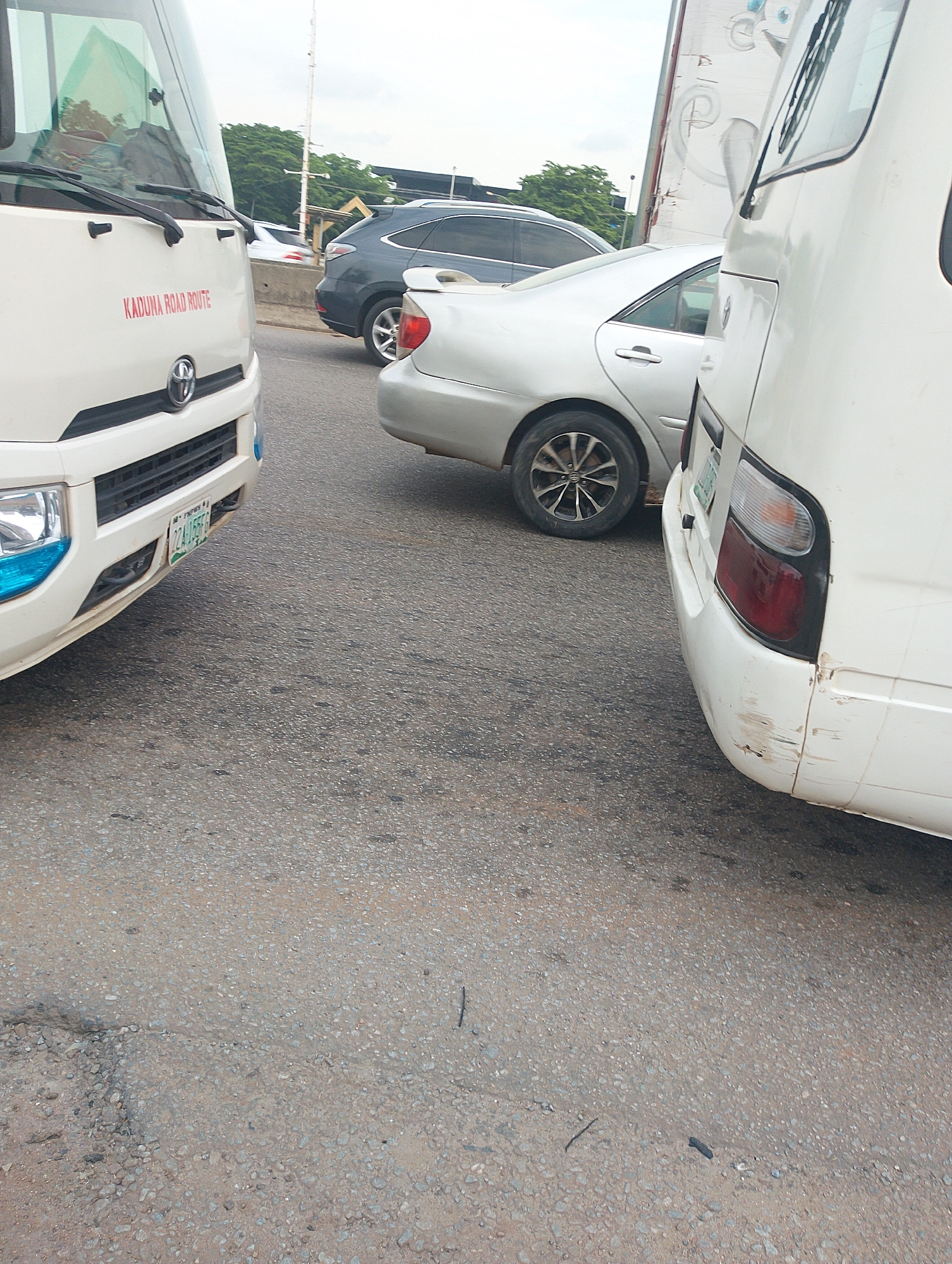 The image depicts a traffic scene on a road in Dutsen Alhaji, Nigeria. The foreground is dominated by the front and side of a white bus, with the text "KADUNA ROAD ROUTE" prominently displayed in red. The bus has a Toyota logo visible on its grille and a license plate that reads "224 JGS 6." The front bumper has blue accents.
To the right of the white bus, another white vehicle, likely a minibus or a van, is partially visible, showing its rear section with taillights and some scuff marks on the bumper.
In the background, a silver sedan is parked behind the white bus. Further back, a dark-colored SUV is also visible, along with lush green trees lining the roadside. Overhead, a utility pole with wires is present. The sky appears overcast, suggesting it might be a cloudy day. The road surface is asphalt, showing signs of wear and tear with small pebbles and cracks. The overall impression is that of a typical traffic situation in a Nigerian urban or suburban area. There are no people visible in the frame.