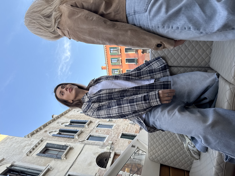 Two individuals are seated within what appears to be a boat, likely a water taxi, on a canal in Venice, Italy. The immediate setting consists of light beige quilted bench seating.
One individual, a woman with long brown hair, is positioned on the left, facing towards the right side of the frame and looking upwards. She wears a white t-shirt, an open blue and beige plaid flannel shirt, and light blue wide-leg jeans. A silver-toned ring is visible on her right ring finger. Her posture suggests she is kneeling or sitting with her knees bent on the bench.
A second individual, visible from the back, is positioned on the right, facing towards the first woman. This person has light blonde or white hair and wears a tan suede-like jacket over light blue jeans. No direct interaction is visible, but their proximity indicates they are together.
The background features historic Venetian architecture. On the left, a tall, multi-story building with cream-colored masonry, multiple windows with dark green shutters, and arched ground-floor features rises directly from the water's edge. Further back and to the right, a vibrant orange-red building with similar window structures and small balconies is visible. The sky above is clear blue with scattered wispy clouds.