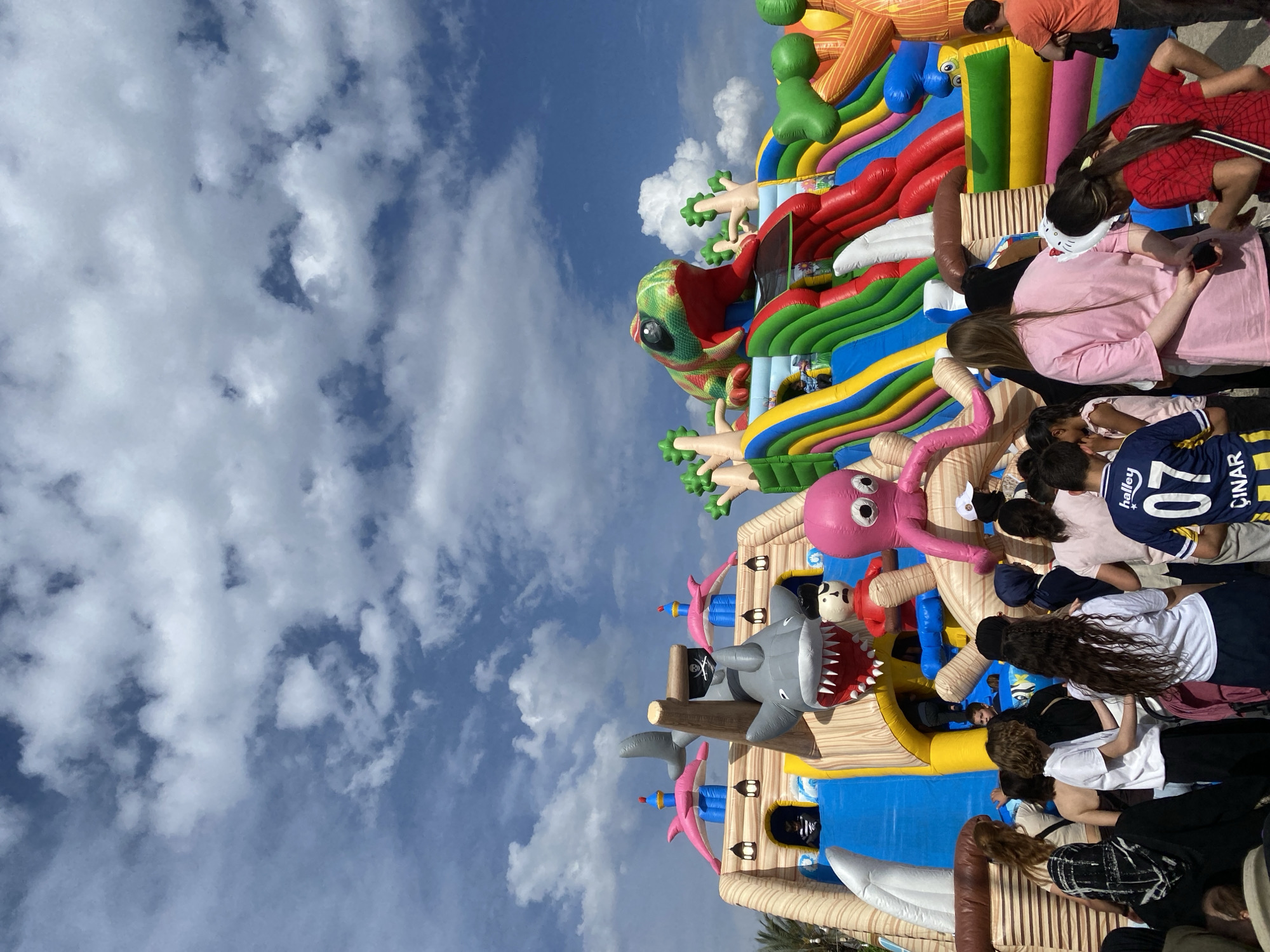 The image shows a crowded inflatable playground on a partly cloudy day, possibly in Alanya, Turkey. The playground features several inflatable structures, including a pirate ship with a shark head, an octopus, and a multi-colored slide with a chameleon at the top. A group of people, mostly children and some adults, are present. They appear to be interacting with the inflatable structures, either climbing or playing on them. Some people are standing nearby. The sky is visible in the background, indicating an outdoor setting.