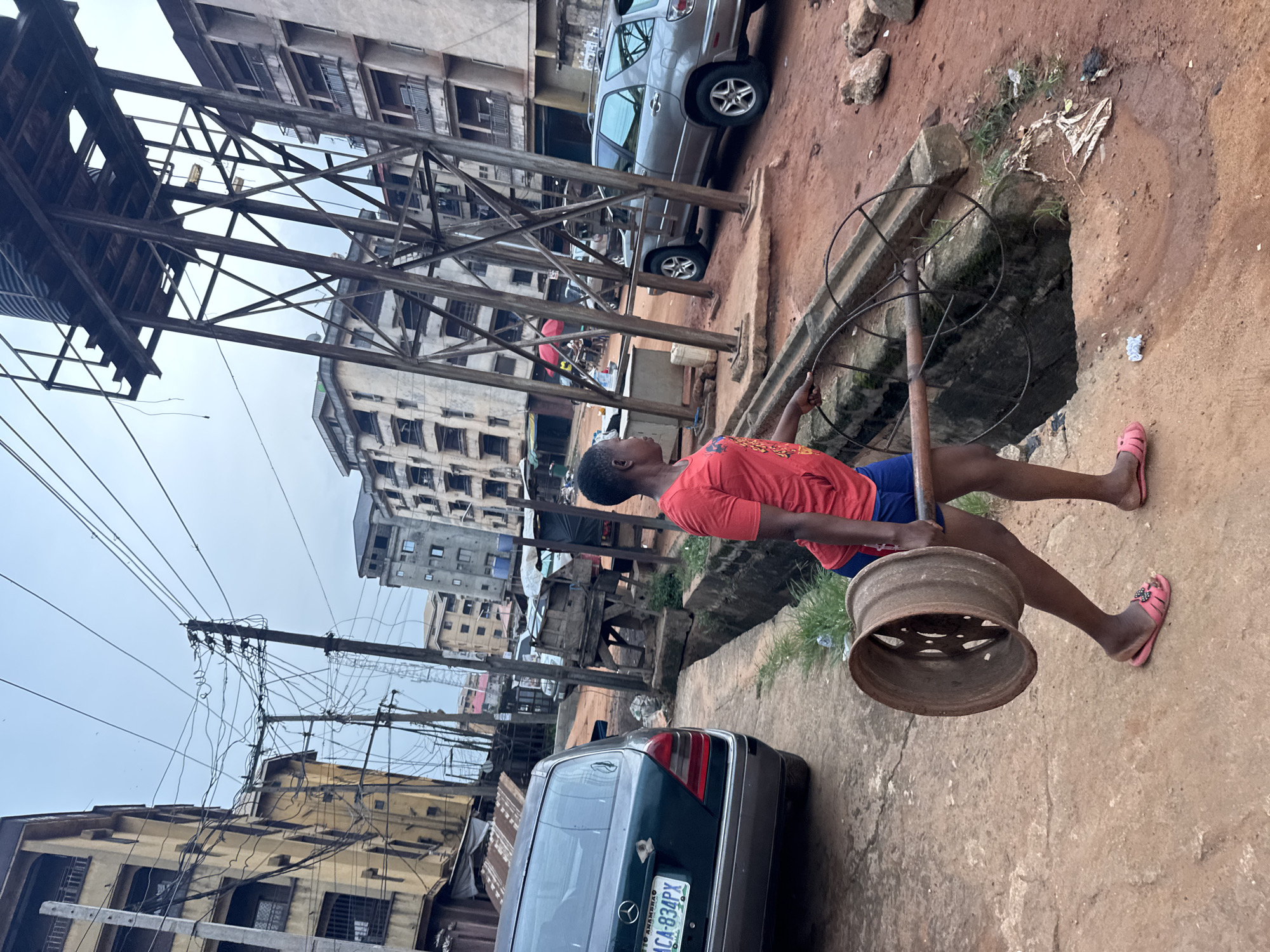 A person is carrying a metal rim along a concrete street in Onitsha, Nigeria. The individual is wearing a red t-shirt, blue shorts, and pink sandals. The street is bordered by buildings on either side, with power lines and utility poles visible overhead. Several parked cars are present. An open sewer is visible on the right side of the street. A license plate is visible on a green vehicle with plate number ACA-834PX Anambra.