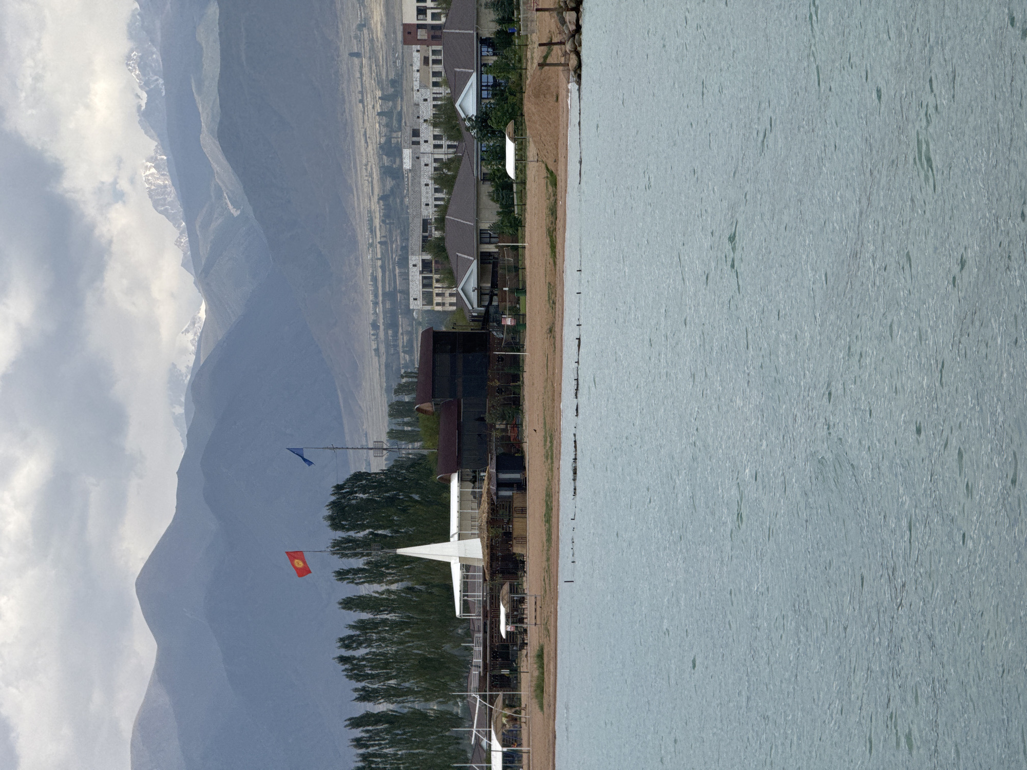 This is an image taken from the water, looking towards a beach area with mountains in the background. The foreground is dominated by the rippling surface of a body of water, likely a lake, with light blue-green hues and visible texture from the waves.
On the shore, there is a sandy beach with various structures. To the left, there are some open-air covered seating areas with thatched roofs, and further back, a tall, white, spire-like structure. In the center and right, there are buildings with dark roofs, some appearing to be restaurants or cafes, with outdoor seating visible. There are also trees lining the edge of the beach and extending into the background.
Behind the beach, there is a town or resort area with several buildings. Some appear to be modern, multi-story structures with many windows, possibly under construction or recently built. There are also some smaller, single-story dwellings with dark roofs closer to the beach.
In the far background, a range of mountains is visible. The lower slopes are dry and sparsely vegetated, while the higher peaks are rugged and appear to have snow caps. The sky is overcast with soft, grey clouds.
Two flags are visible on flagpoles in front of the trees: a red flag with a yellow emblem (likely the flag of Kyrgyzstan) and a blue flag. The overall scene suggests a resort or recreational area on the shore of a lake, with a backdrop of dramatic mountain scenery. The location is identified as Sary-Oy, Kyrgyzstan. There are no people clearly visible in the image.