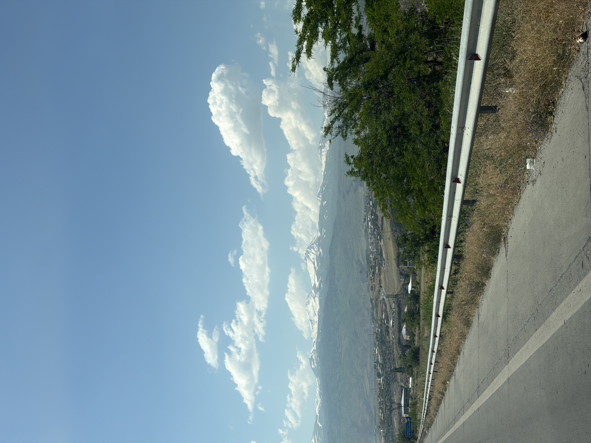 The image depicts a scenic view of the Aragats mountain range, as seen from a road in Ashtarak, Armenia. The foreground features a paved road with a white line marking its edge, bordered by dry grass and a metal guardrail. Beyond the road, a valley unfolds with scattered houses and a winding road, leading towards the majestic mountains in the distance. The Aragats mountains are snow-capped, indicating a significant altitude. The sky is a clear blue with scattered white clouds, suggesting a sunny day. A few green trees are visible on the right side of the image, framing the view. There are no people or animals visible in the frame.