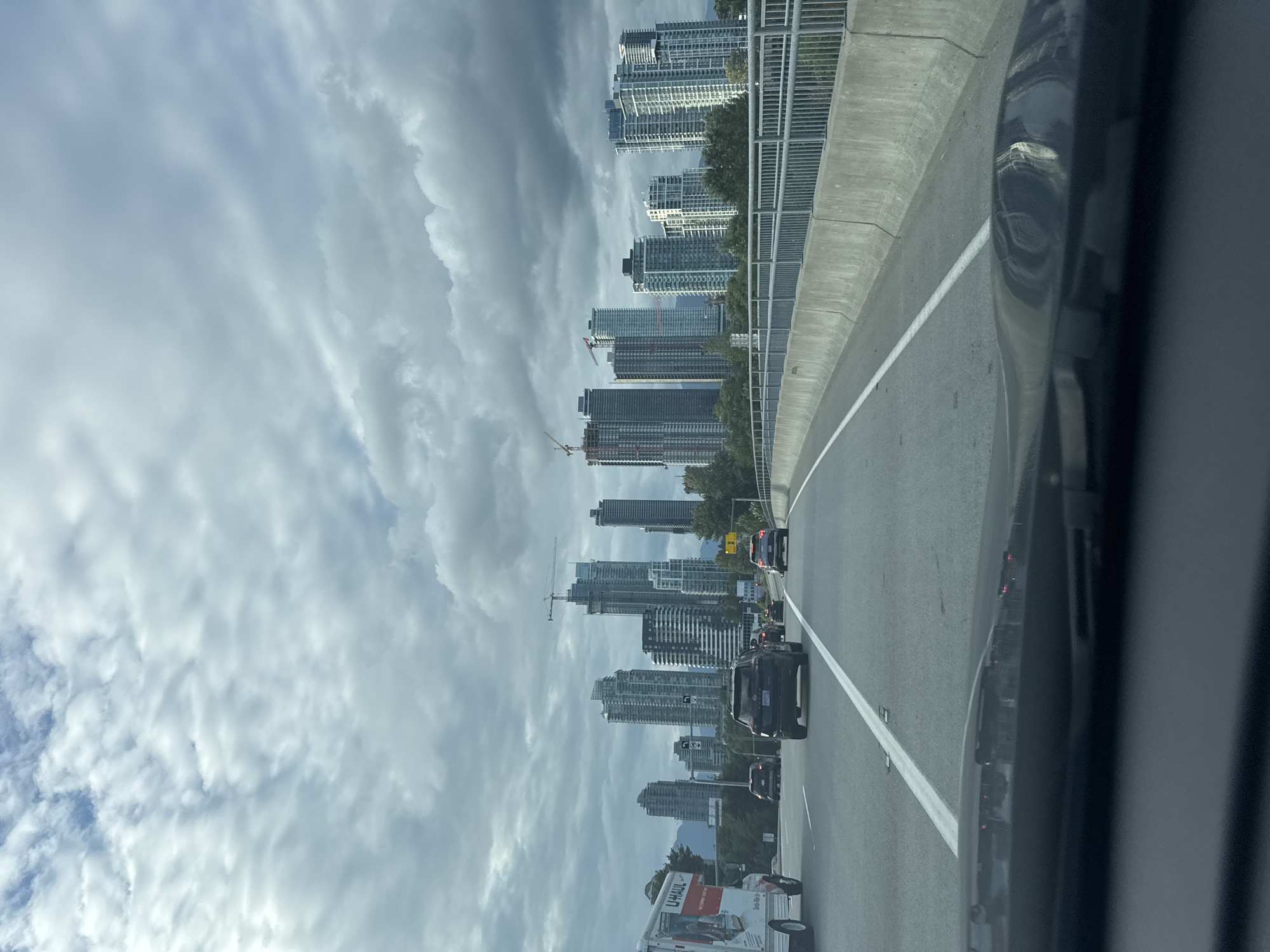 The image depicts a view from a vehicle driving on a multi-lane highway towards a dense urban skyline under a cloudy sky. The highway has clear white lane markings and a concrete barrier with a metal railing on the right side. Several vehicles are visible on the highway, including a U-Haul truck on the left, and other cars and SUVs in front.
In the background, a modern cityscape characterized by numerous tall residential or commercial buildings is prominent. Several of these buildings appear to be under construction, with cranes visible. The skyline suggests a significant metropolitan area.
Based on the visual cues, particularly the characteristic architecture and the presence of a U-Haul truck, the location is identified as Burnaby, Canada, likely on a major arterial route such as Highway 1 (Trans-Canada Highway) or Boundary Road, as it approaches the Metrotown area or the False Creek/Downtown Vancouver skyline. The environment is urban, with the highway cutting through the landscape and leading into the heart of the city. The cloudy sky indicates overcast weather conditions.