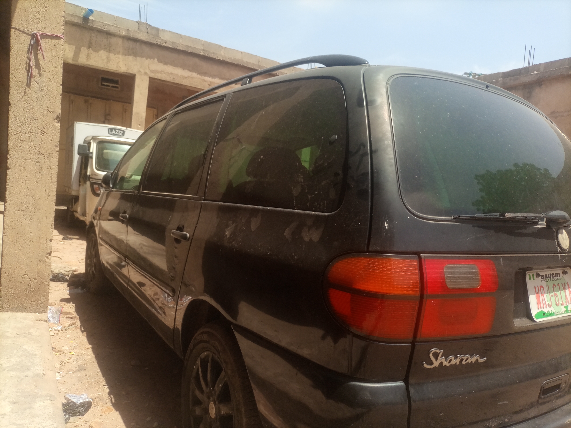 In Arawa, Nigeria, a black Volkswagen Sharan minivan is parked on a dirt lot next to a building. The minivan is facing away from the camera, and its rear is visible. The license plate is from Bauchi, Nigeria, and reads "NRJ 61XA". A white truck with the word "LAZIZ" on its side is parked in front of the minivan. The building appears to be made of concrete blocks and has a corrugated metal roof. The ground is covered in dirt and debris. The sky is clear and blue.