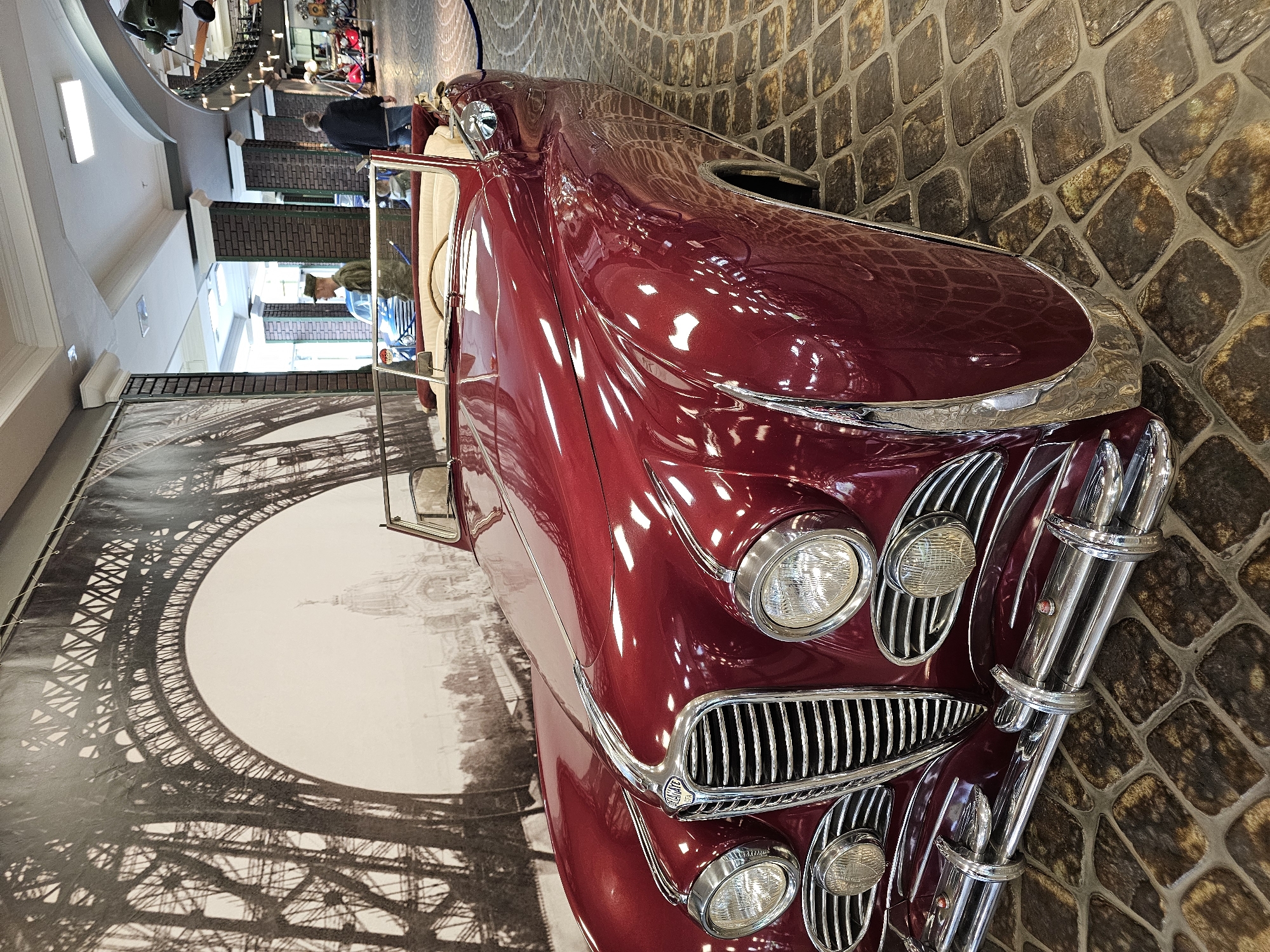 A high-angle shot shows the front of a maroon vintage car angled in front of a black and white backdrop of the Eiffel Tower. The car has a chrome grill and headlights and is parked on a cobblestone surface. In the background, people stand near a railing in a brightly lit museum setting. This image was taken in Архангельское, Russia.