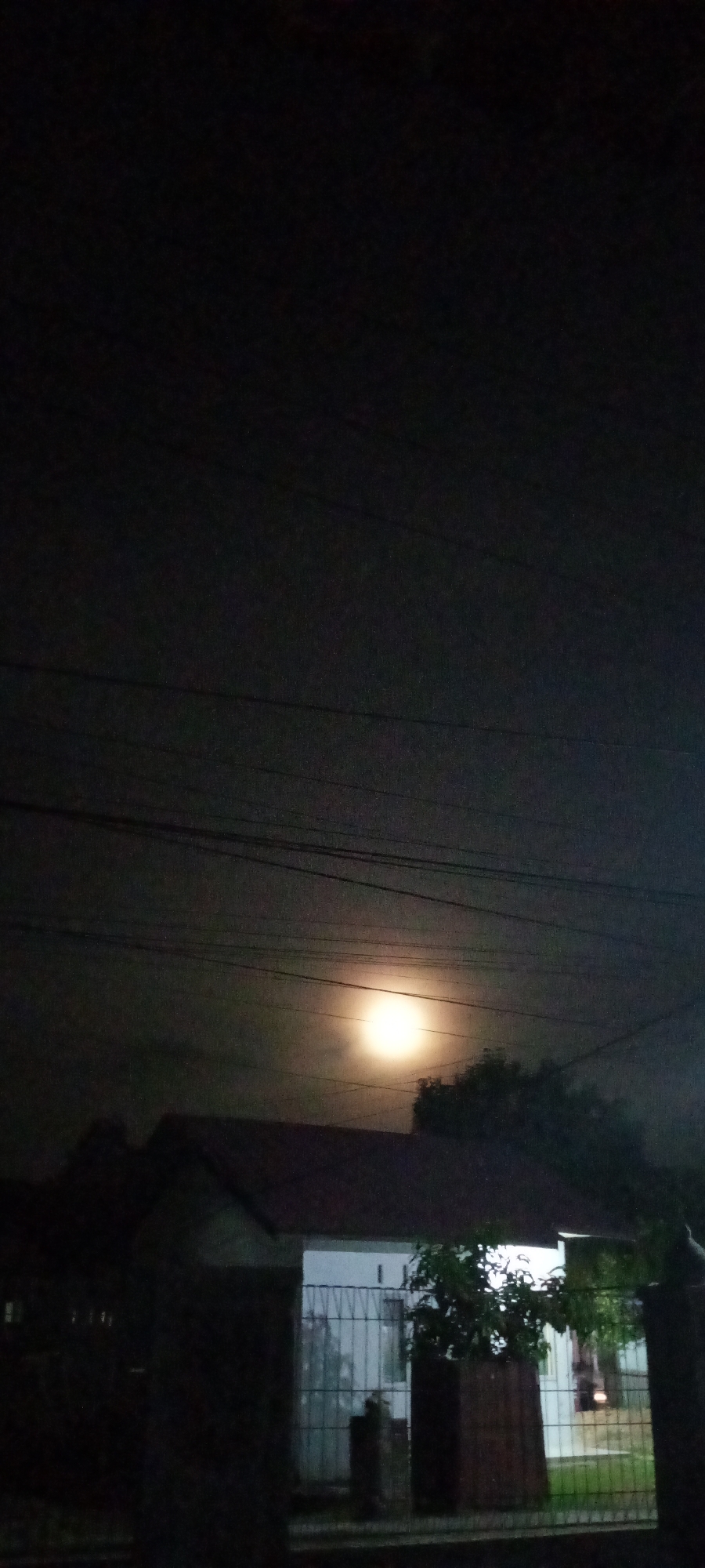 Captured at night, the image depicts a street scene in Lhokseumawe, Indonesia. In the foreground is a metal fence, partially obscuring a white, single-story house with a red tile roof. A small tree or bush is planted near the fence, its leaves partially visible in the dim light. Overhead, power lines stretch across the sky. In the background, a bright moon or streetlamp illuminates the scene. The sky is dark with some clouds visible. The overall ambiance suggests a residential area at nighttime. No people are visible in the shot.
