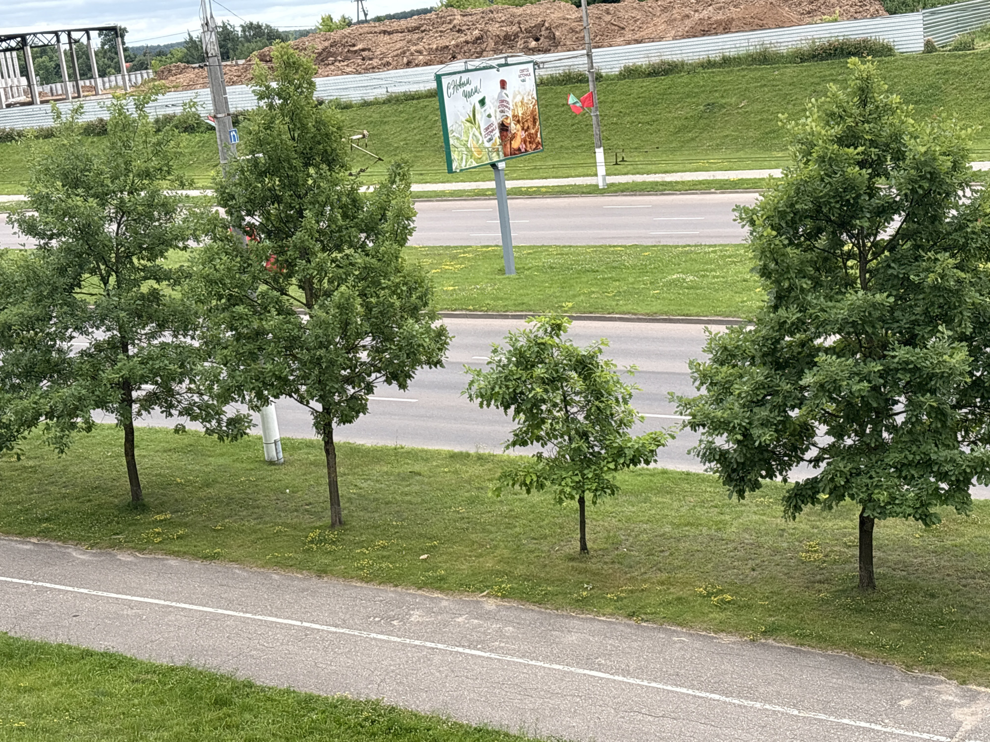 The image shows a roadside scene in Vitsebsk, Belarus, under a cloudy sky. In the foreground, a paved pathway with a white line runs parallel to a grassy area. Beyond the pathway and grass, a road with traffic lanes is visible. Lining the road are several deciduous trees with full green foliage. In the middle ground, a billboard advertising beverages, likely tea or juice, is mounted on a pole. Two small flags, one red and one red and green, are attached to the pole next to the billboard. Behind the road and billboard, a sloped, grassy embankment rises, topped by a large mound of reddish-brown material, possibly construction debris or earth. In the far background, structures that appear to be part of a construction site are visible, including metal framing and fencing. The overall environment suggests an urban or suburban setting during daytime.