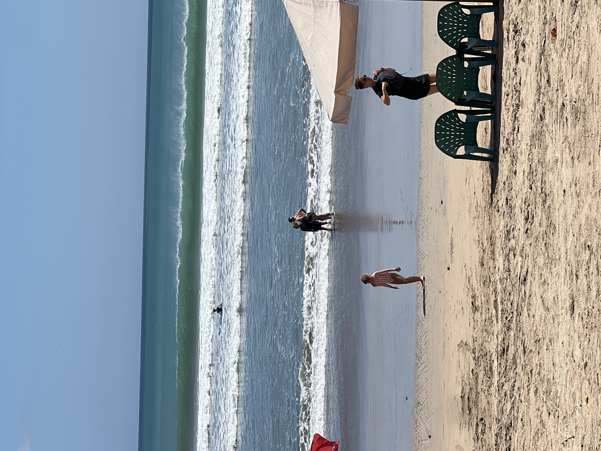 The image captures a sunny day at Kuta Beach, Indonesia, characterized by its wide sandy shore and consistent waves. In the foreground, three green plastic chairs are arranged on the sand, partially shaded by a light-colored beach umbrella. A woman in a black dress stands near the umbrella, adjusting her backpack. Further back, a young girl in a pink and white striped swimsuit walks on the wet sand. In the shallow water, a group of three people stand together. Beyond them, several surfers are visible in the ocean, with waves rolling in. The horizon shows a clear blue sky meeting the turquoise ocean.