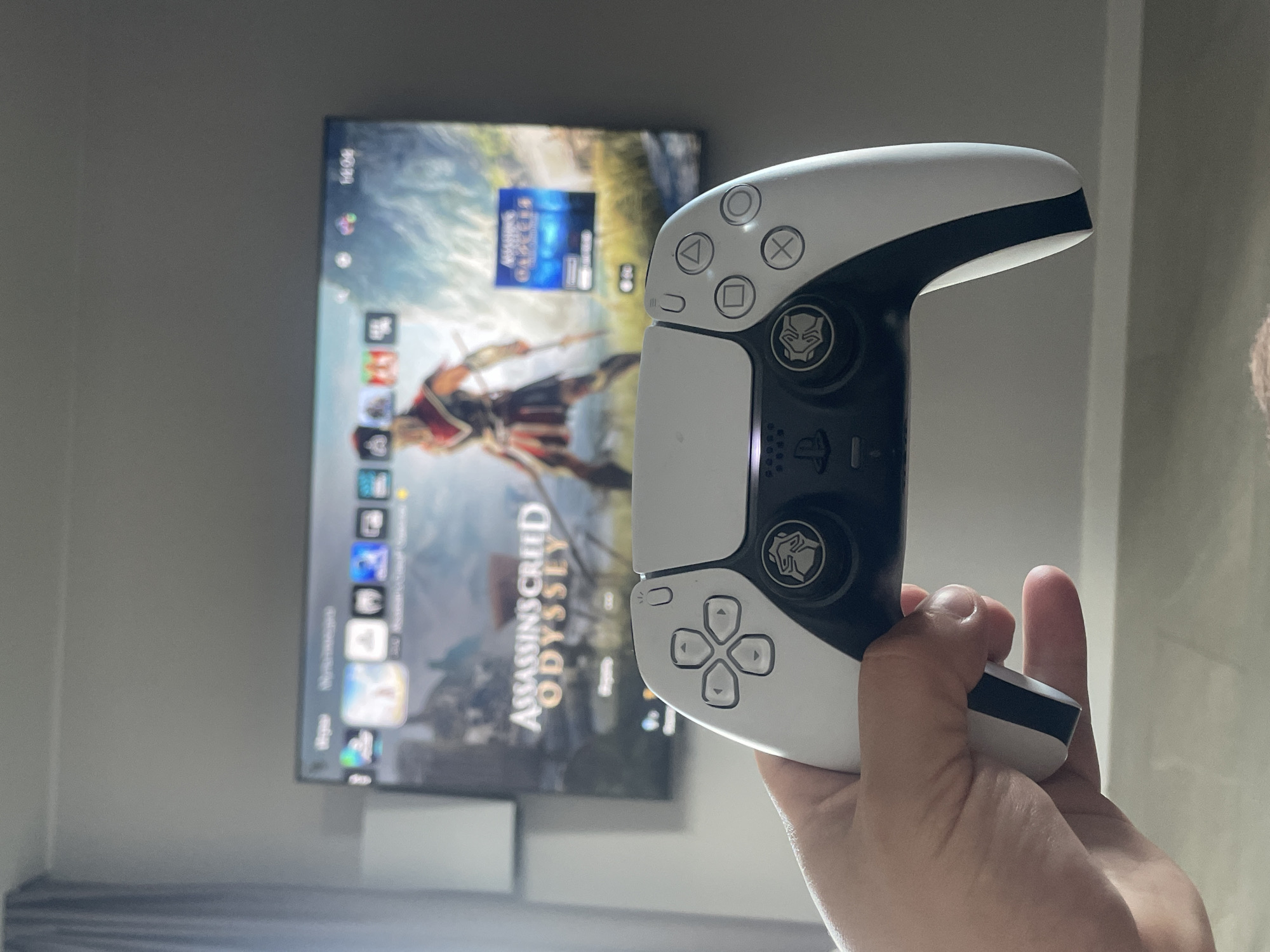 A person is holding a PlayStation 5 controller in their right hand. The controller is white and black and has custom thumb sticks featuring the Marvel Black Panther character logo. Behind the controller, a television screen displays the PlayStation 5 user interface, with the game "Assassin's Creed Odyssey" highlighted. The time is shown as 16:06. The location is not Эстачи, Russia, as the provided image does not contain enough contextual information to determine the location.