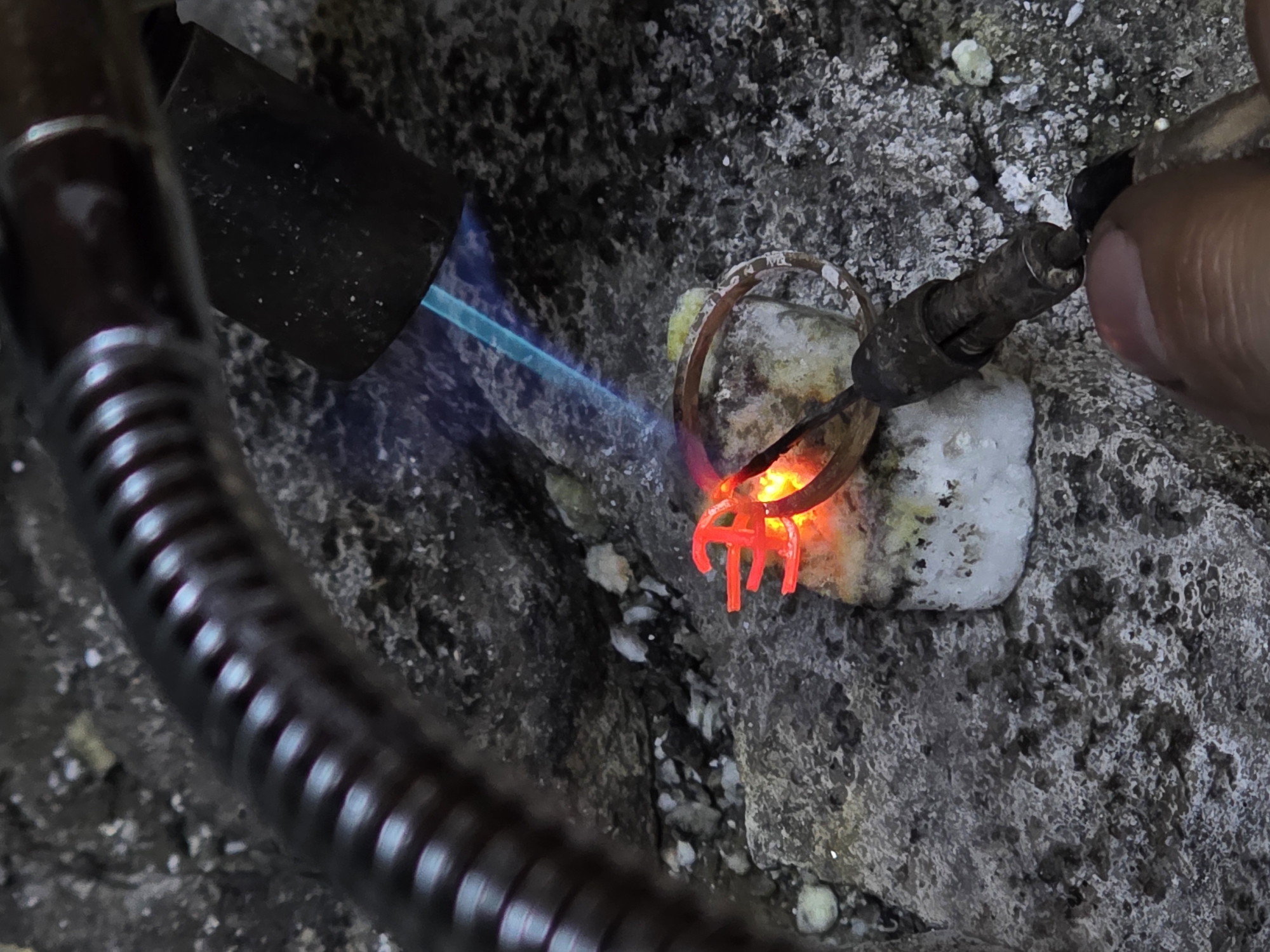 The image depicts a close-up view of a close-up of a soldering process, where a small object with a '€' symbol, glowing red from heat, is being held by a tool. A torch flame, emitting blue light, is directed towards the object and a metal ring. The scene appears to be outdoors, with a rocky, textured surface in the background, possibly representing a natural environment or an industrial setting. No specific location in Kazan, Russia is identifiable from the visual information provided.