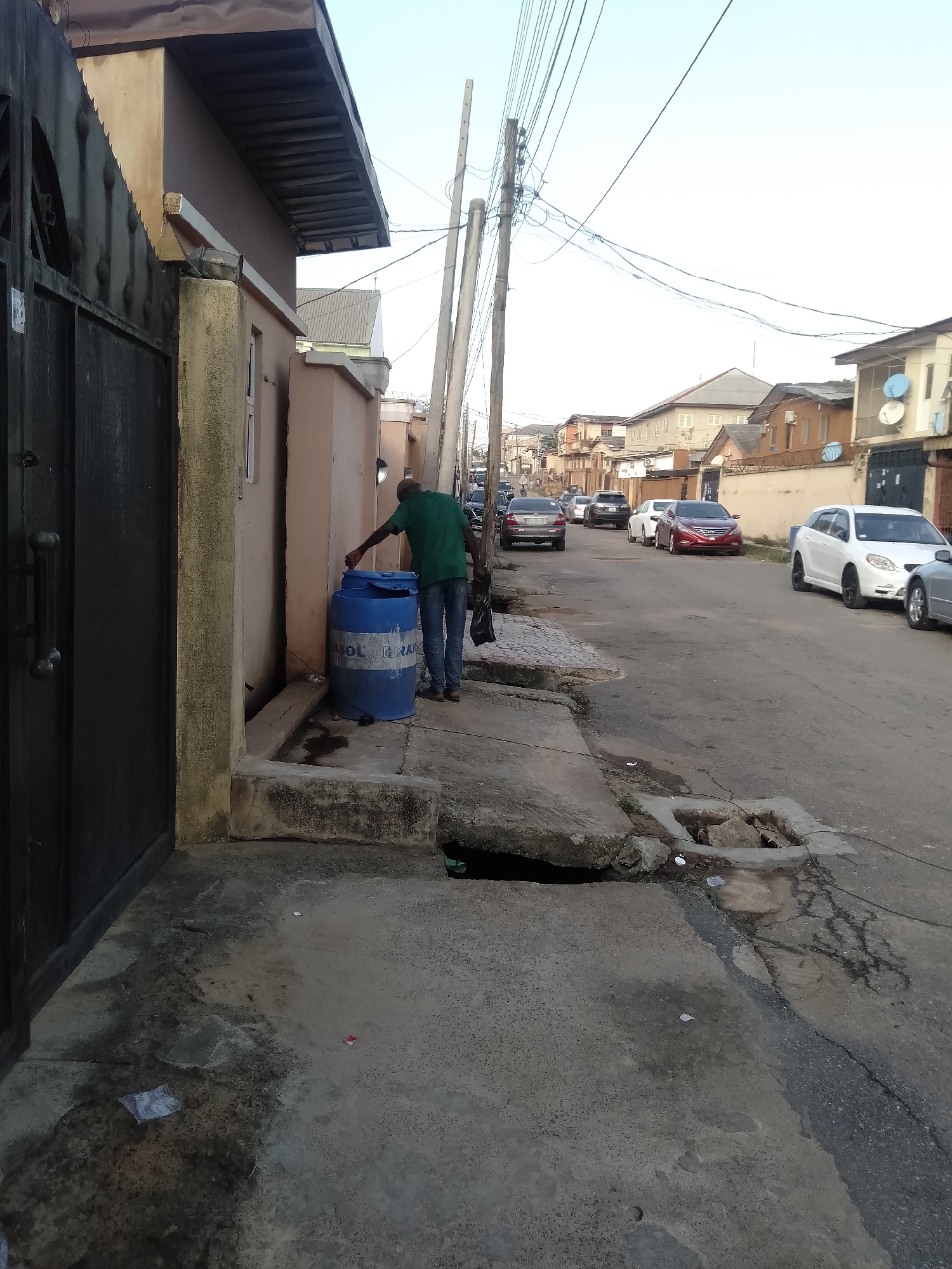 An eye-level outdoor shot depicts a residential street scene, likely in Shangisha, Nigeria, during what appears to be late afternoon or early morning, given the soft, bright light and clear sky.
In the foreground to the left, a dark metal gate belonging to a building is partially visible. Adjacent to the gate, a man wearing a green t-shirt and blue jeans is seen from behind, attending to a large blue plastic barrel with a lid. Partial text "NAOL" and "GRAS" is visible on the barrel's side. The barrel sits on a concrete pavement, which shows signs of use and some damp patches.
To the right of the man, the concrete pavement continues, featuring an open drain or gutter with debris. Utility poles with a dense network of overhead wires line the street, extending into the background. The street itself is paved and stretches into the distance, with several cars parked along both sides. Vehicles include a white wagon-style car, a red sedan, a grey sedan, and other darker sedans and SUVs.
The street is flanked by various residential buildings of different heights and colors, some with satellite dishes visible on their facades. The architecture suggests a local urban or suburban setting. The overall scene captures a slice of daily life in a bustling neighborhood. Some litter, such as a plastic bag and a small white cup, is visible on the ground.