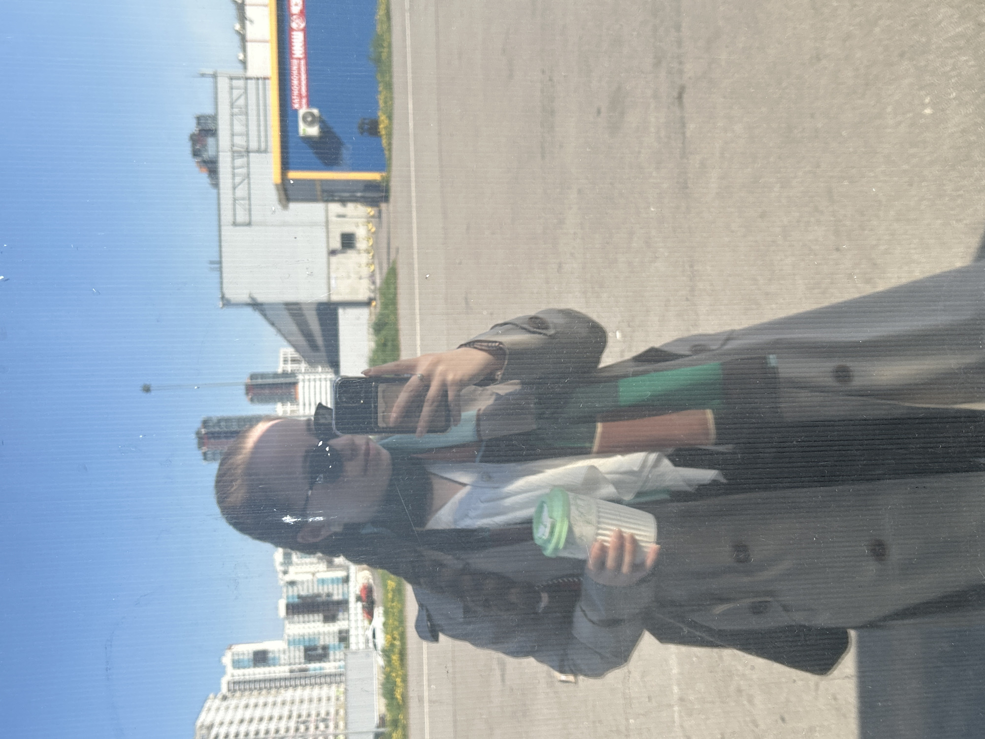 The image shows a reflection in a glass surface, likely a window. The reflection depicts a person wearing sunglasses and a trench coat, holding a reusable coffee cup. The person appears to be taking a photo with their phone, which is visible in the reflection.
In the background, reflected in the glass, are urban buildings of varying heights, indicating a city environment. The sky is blue and clear. Based on the architecture and overall appearance, the city is Saint Petersburg, Russia. The buildings are a mix of older and more modern structures, typical of the city's diverse architectural landscape. A highway passes in front of the buildings and beside the person.