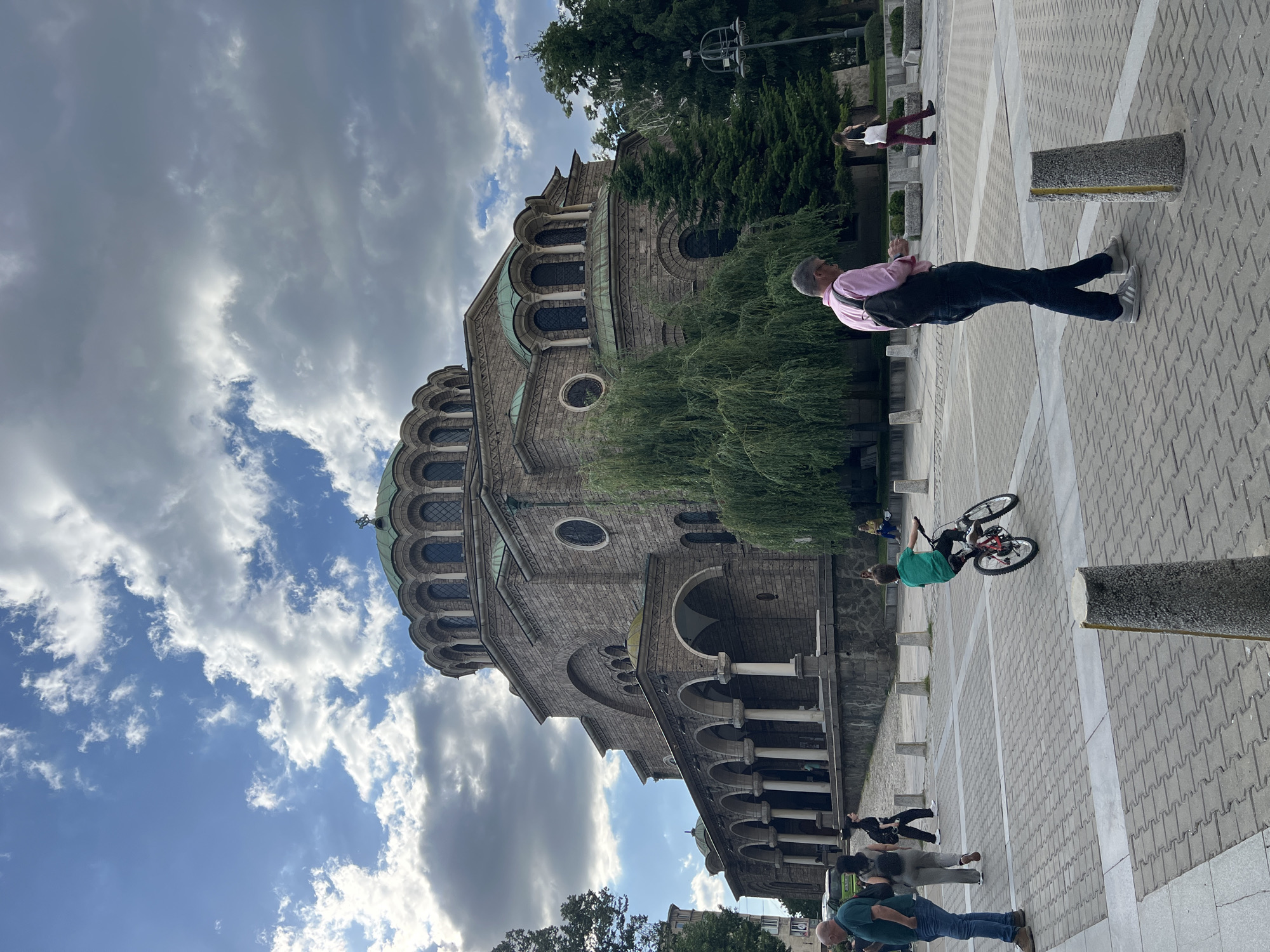 The image captures a scene in Sofia, Bulgaria, featuring the Alexander Nevsky Cathedral. The cathedral, built of grey stone, is partially visible from a low angle, emphasizing its architectural details such as domes and arched windows. The sky above the cathedral is cloudy, with patches of blue visible. In the foreground, a paved area is visible, with several people present. A man in a pink shirt and dark jeans stands near the cathedral, while another person is riding a bicycle. Other people are milling around in the background. The image gives a sense of a public space with people engaging in everyday activities.