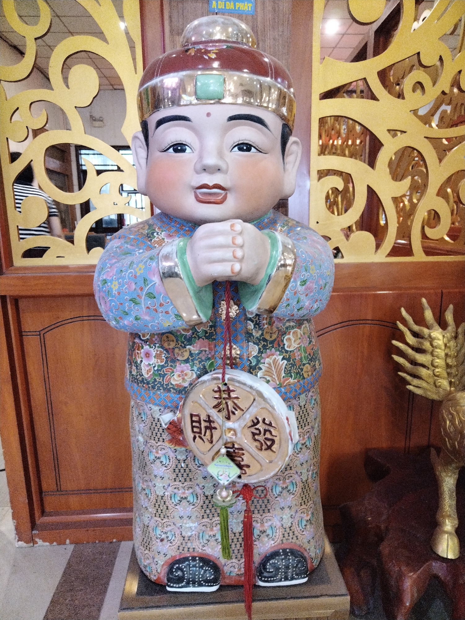 In Chu Văn An Ward, Vietnam, a ceramic statue of a child-like figure stands on a platform. The figure is dressed in a patterned robe with floral designs and a high collar. Its hands are clasped together in front. It wears a red cap with a gold band and a green embellishment. Hanging from the statue's neck is a circular pendant with Chinese characters. Behind the statue are dark wooden panels and a decorative gold screen with swirling patterns. To the right of the statue, a golden, stylized animal figure is partially visible. The scene is indoors, likely in a temple or a place of worship.